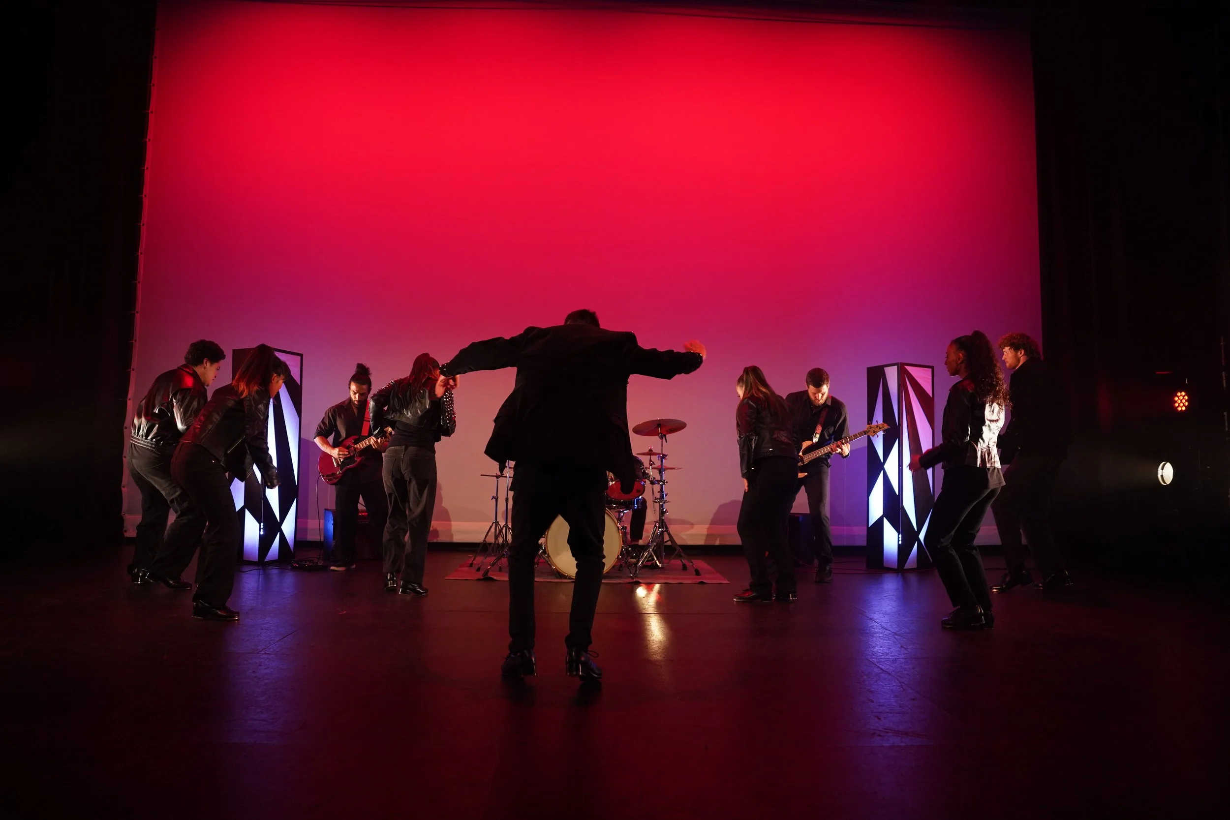 A group of people on stage performing with musical instruments, pink and purple lighting, and a large blank screen in the background.