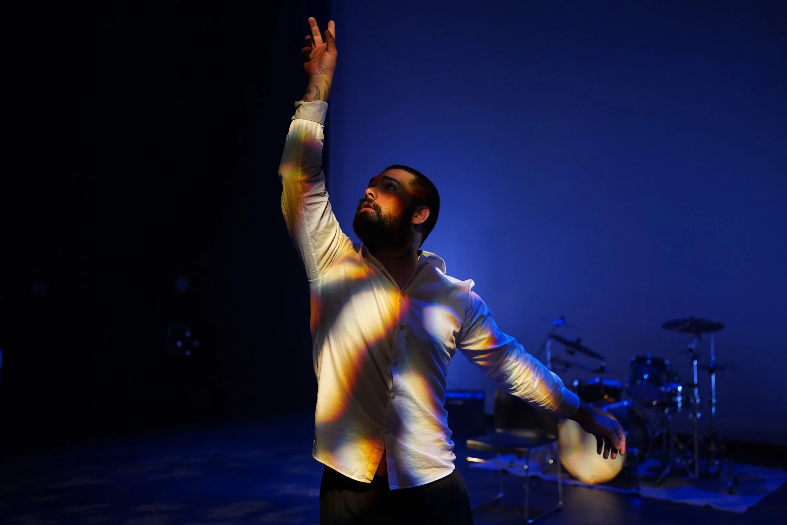 A man with a beard and tattoos, dressed in a white shirt, is raising his right hand with two fingers extended against a dark blue background. There are drum equipment and guitars partly visible in the background, indicating a music or performance setting.