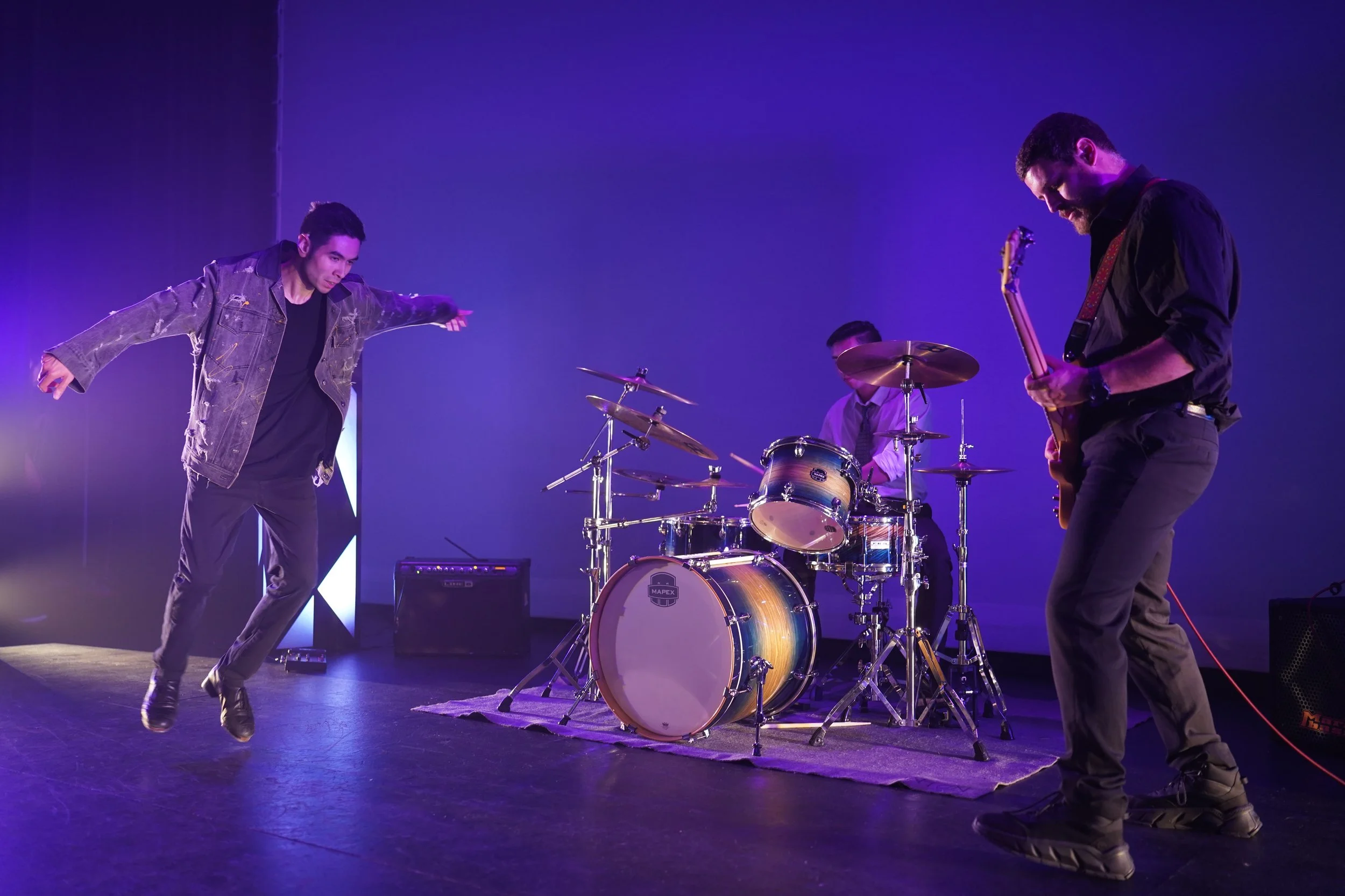 Three musicians performing on stage: one is jumping with arms outstretched, one is playing the drums, and the other is playing the guitar. The stage has purple lighting and a blue background.