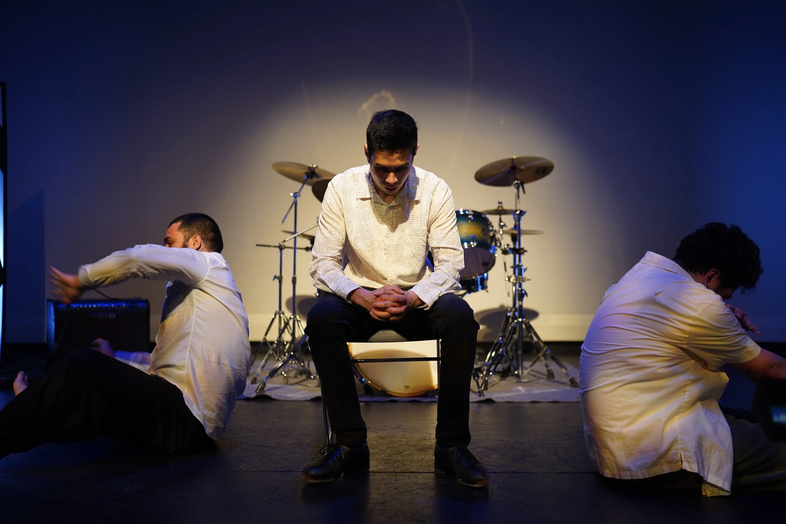 Three performers are on stage performing a dance with a drum set in the background, and the lighting forms a halo effect around the central performer.