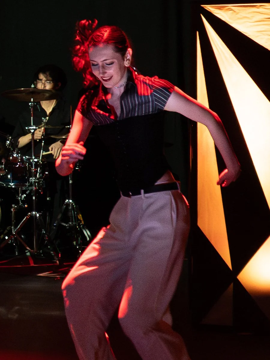 A woman with a ponytail, wearing earrings, a necklace, and a black and white stripped shirt, is smiling and standing with her arms near a drum set. The background is dark.