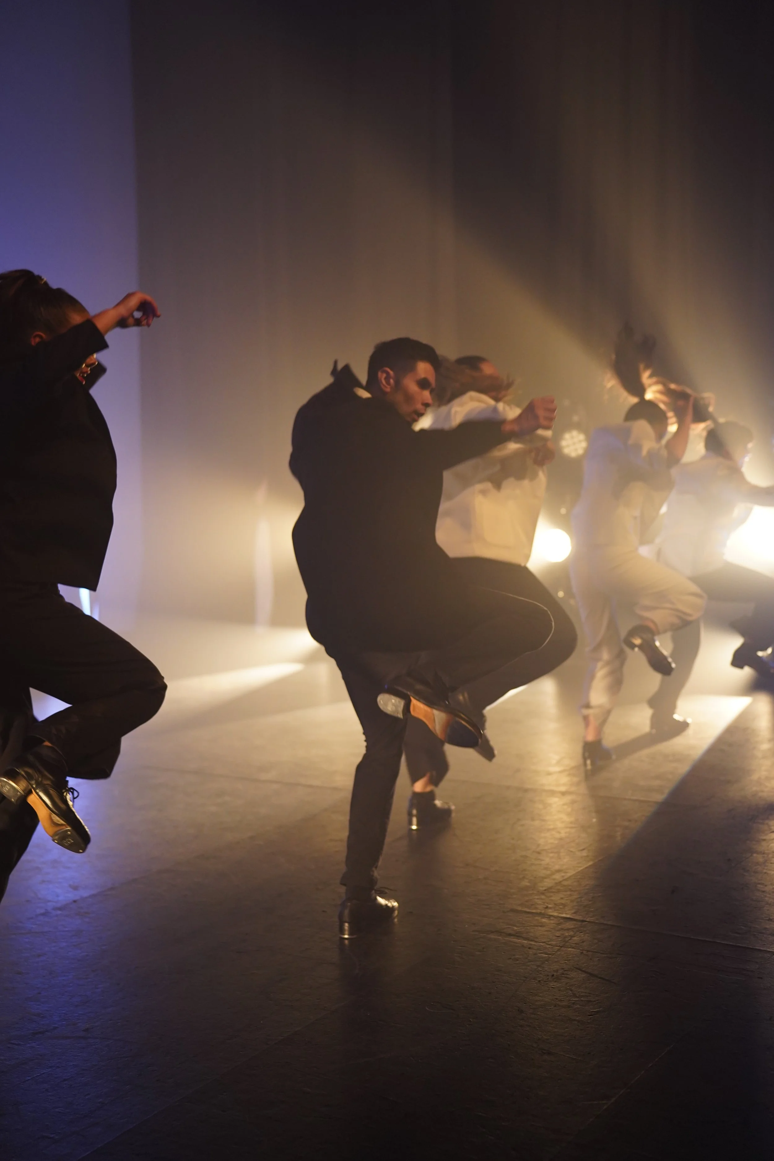 A group of dancers performing on stage, dressed in white and black clothing, with some wearing high heels and moving in sync with raised knees and arm movements, under dramatic lighting and fog effects.