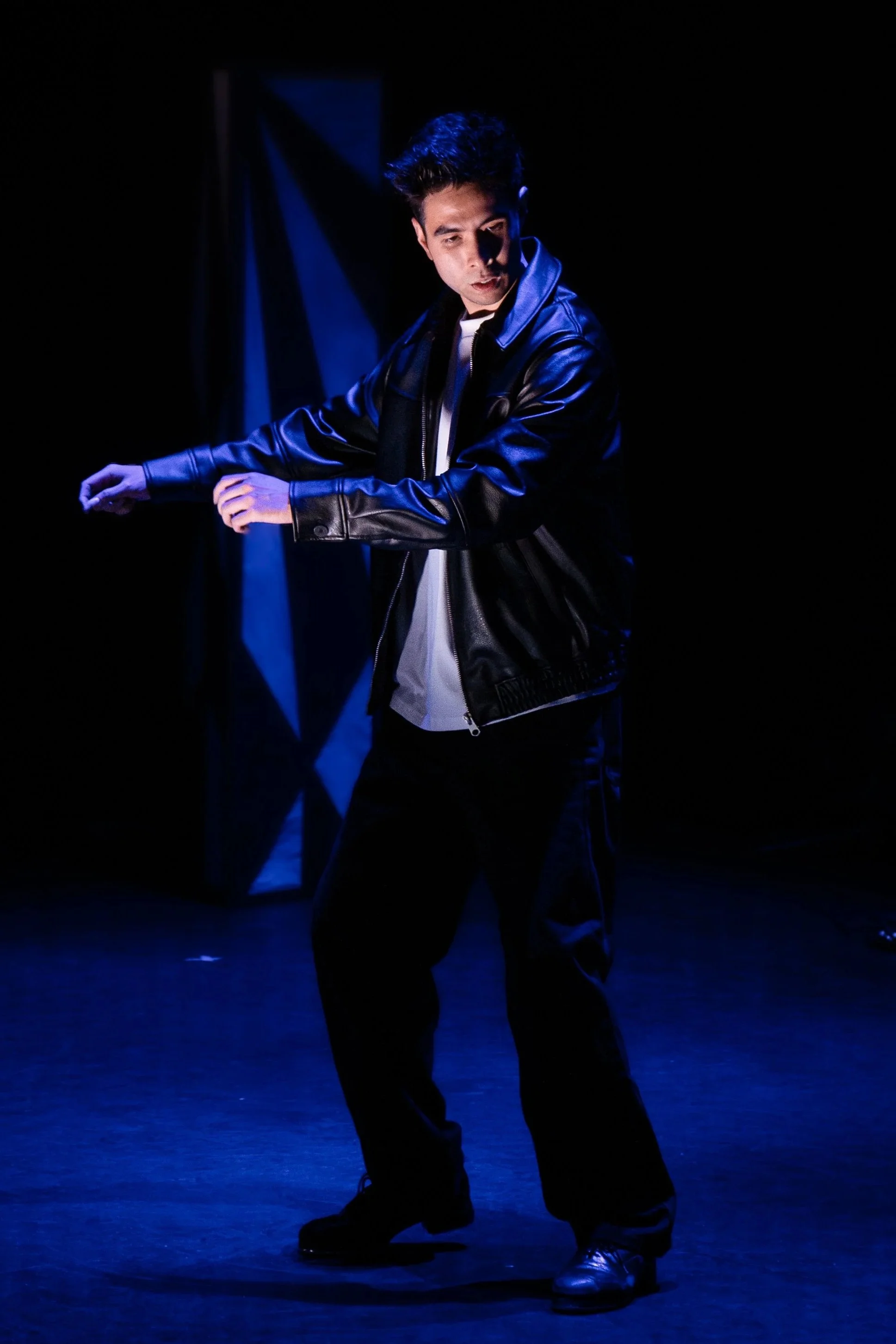 A young man dancing on a dark stage illuminated by blue lighting, wearing a black leather jacket, black pants, and black shoes.