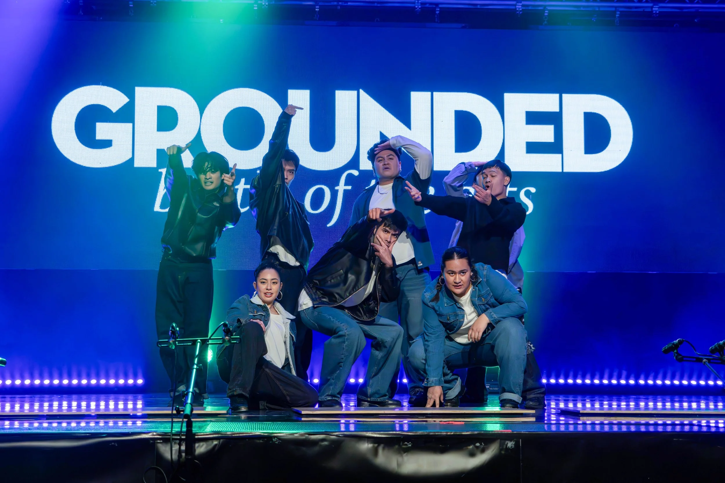 A group of seven young performers on stage, posing in front of a large screen with the word 'GROUNDED' displayed. The stage is lit with colorful lights, and the performers are dressed in casual clothing, striking expressive poses.