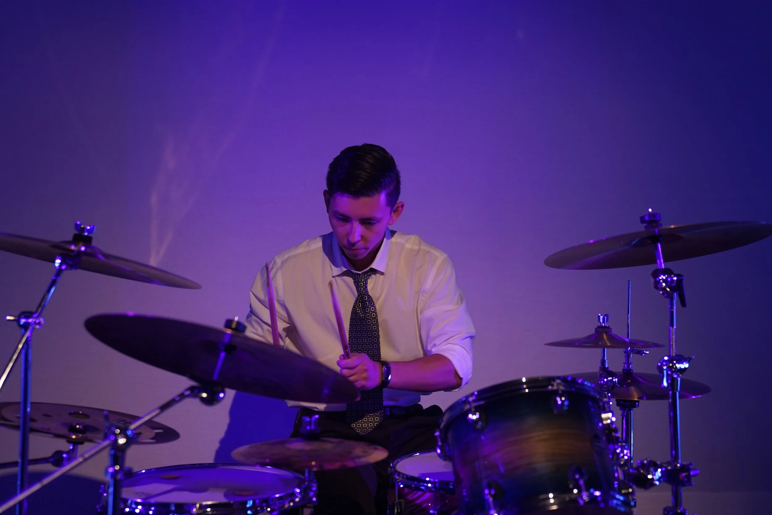 A man in a white shirt and patterned tie playing drums against a purple backdrop.