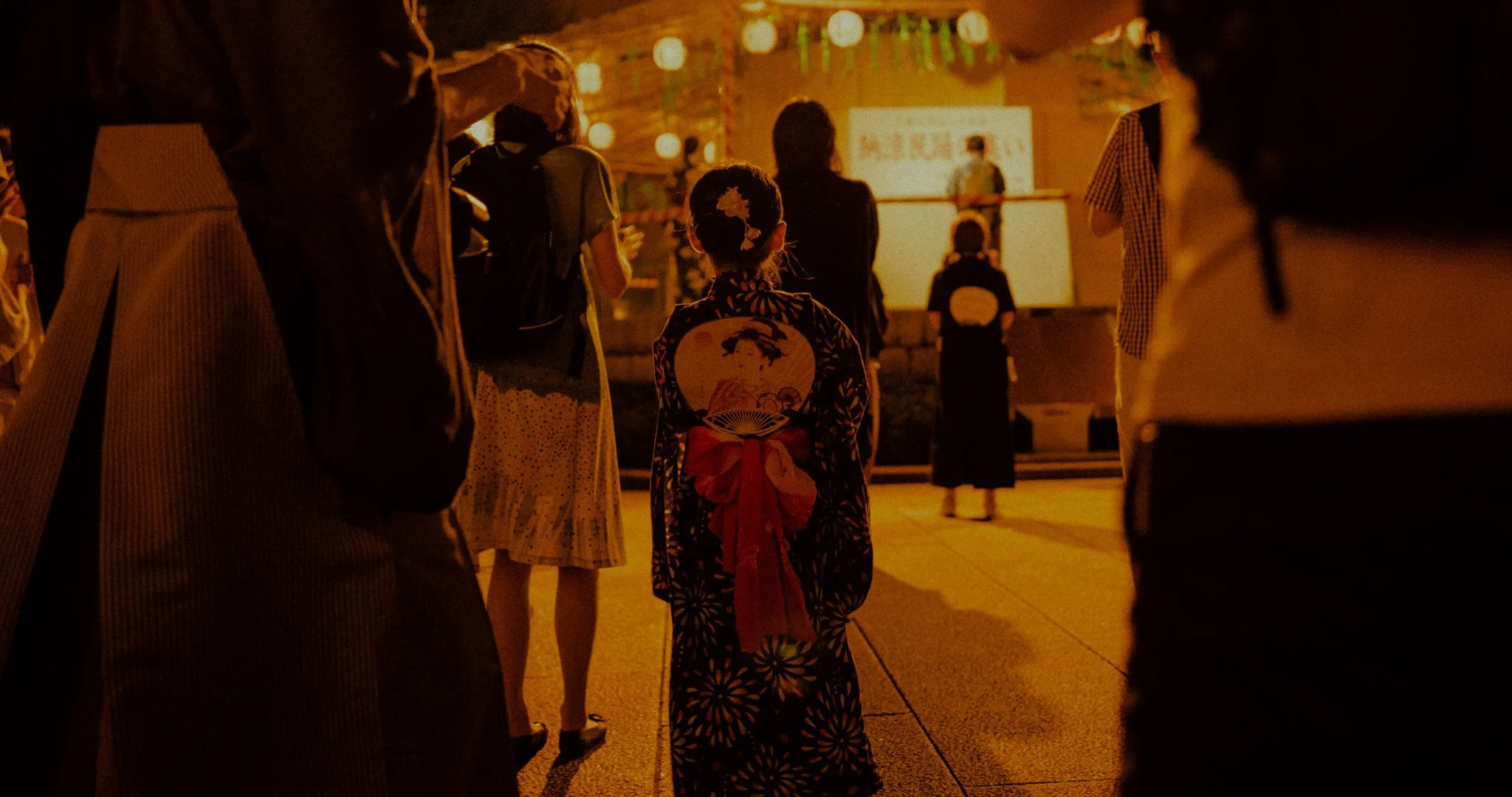 She stands where the crowd thins, as if waiting for someone who will not arrive.