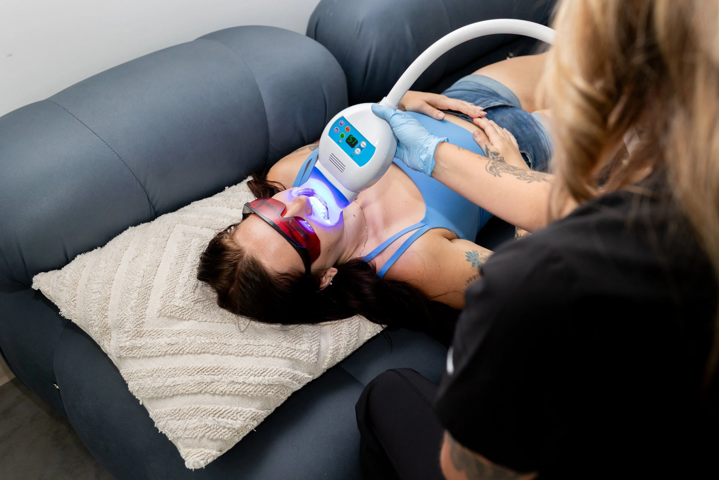 A woman receiving a dental procedure with an LED light and tools, lying on a couch with a towel, wearing protective glasses, and a person with gloves attending to her.
