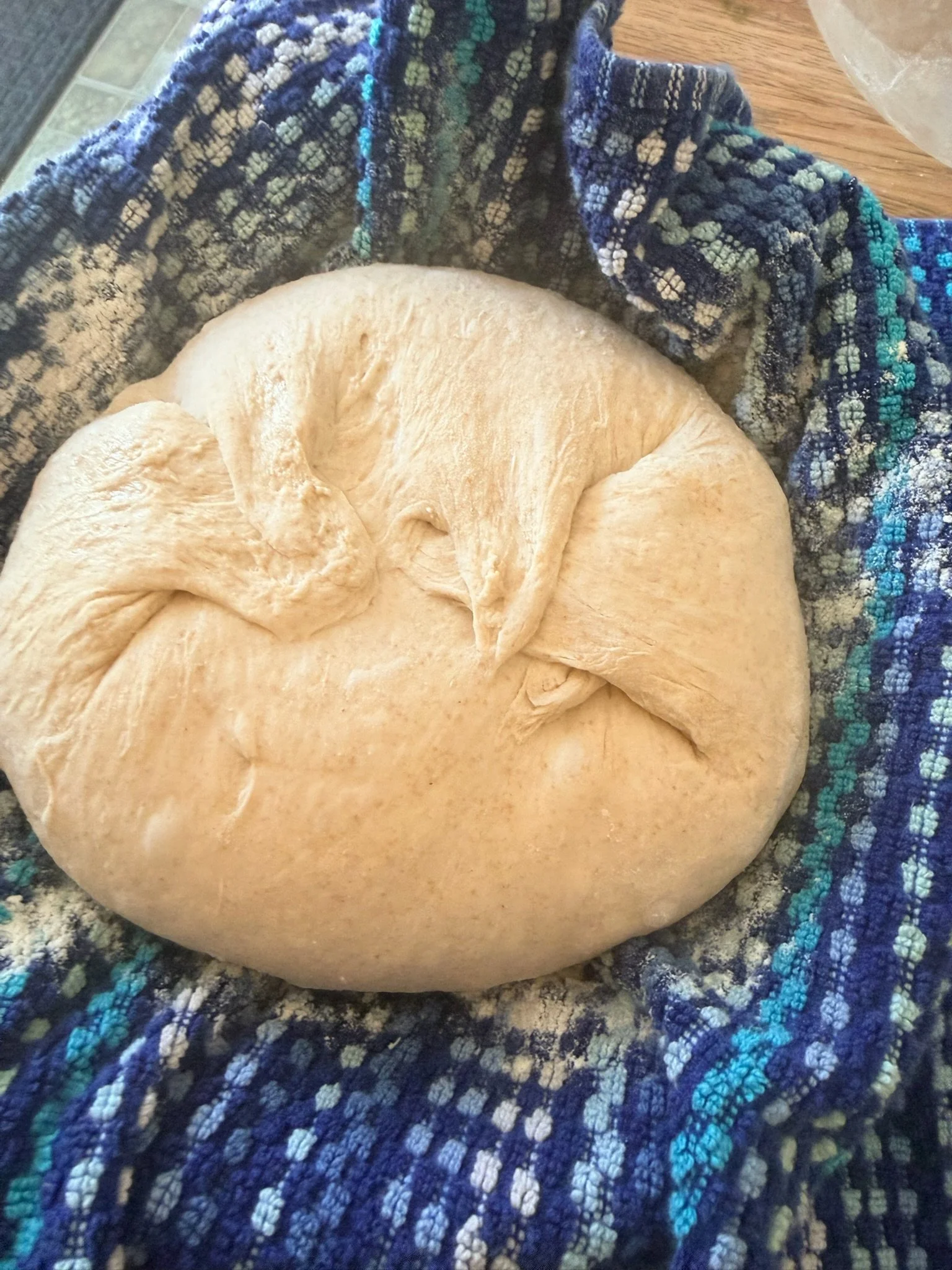 A round loaf of bread resting on a blue patterned cloth with a wooden surface nearby.