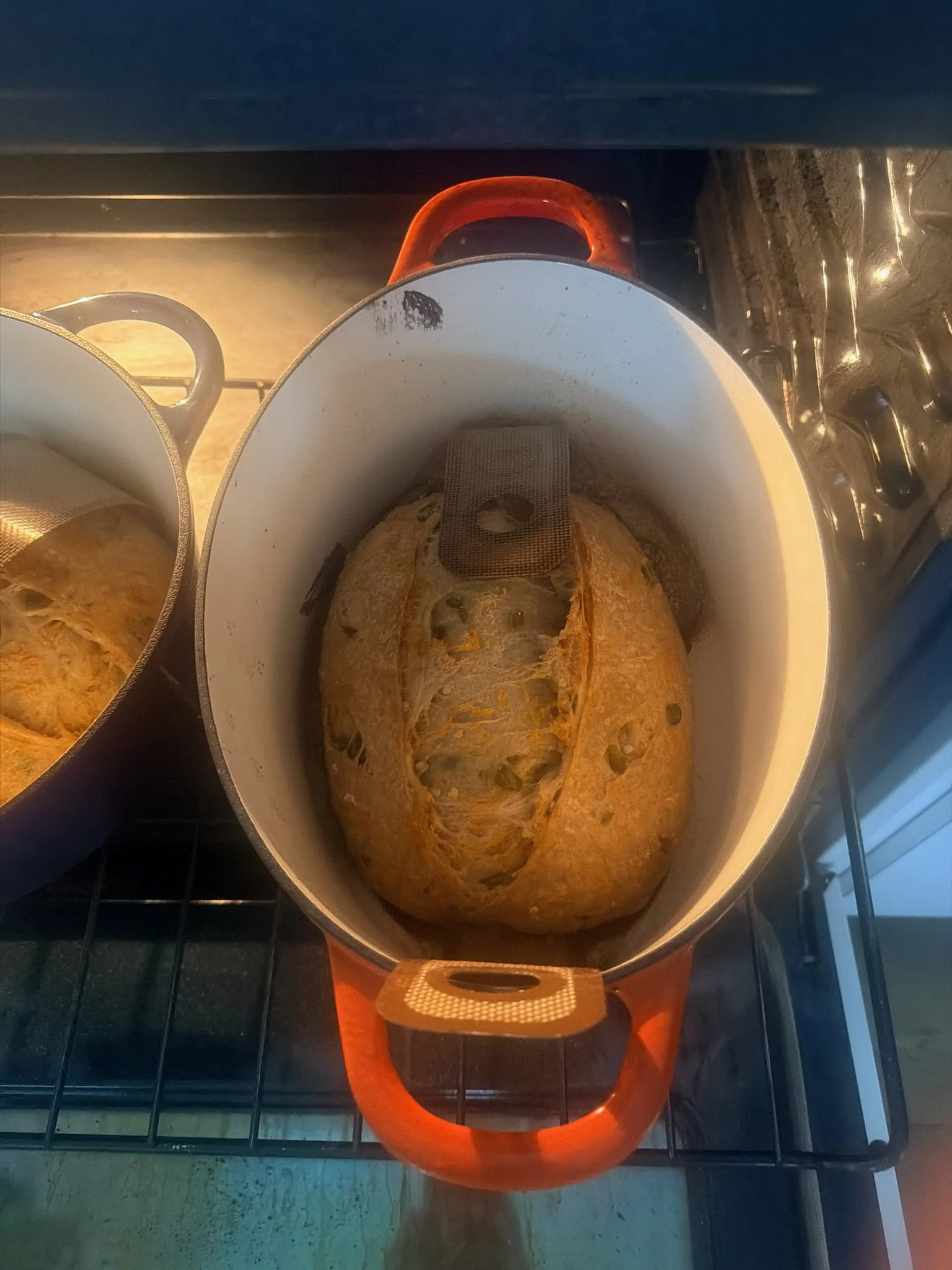 A loaf of bread inside an orange Dutch oven on a stove, with part of another similar loaf visible to the left.