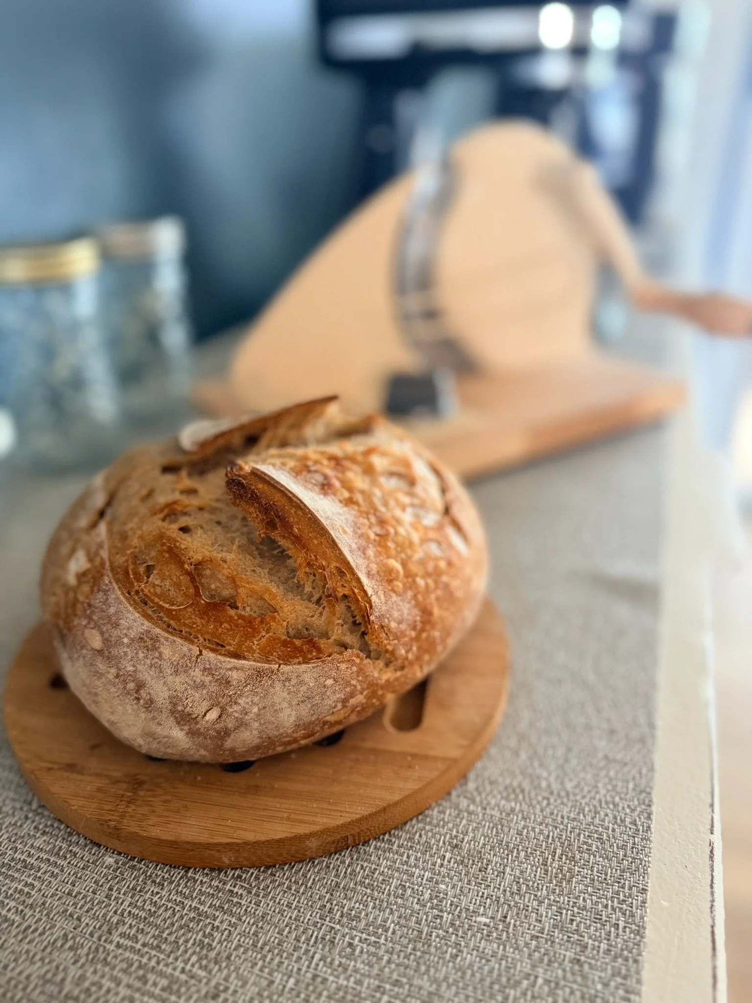 A loaf of crusty bread on a wooden cutting board, with a bread knife, placed on a textured gray surface, and a blurry kitchen scene in the background.
