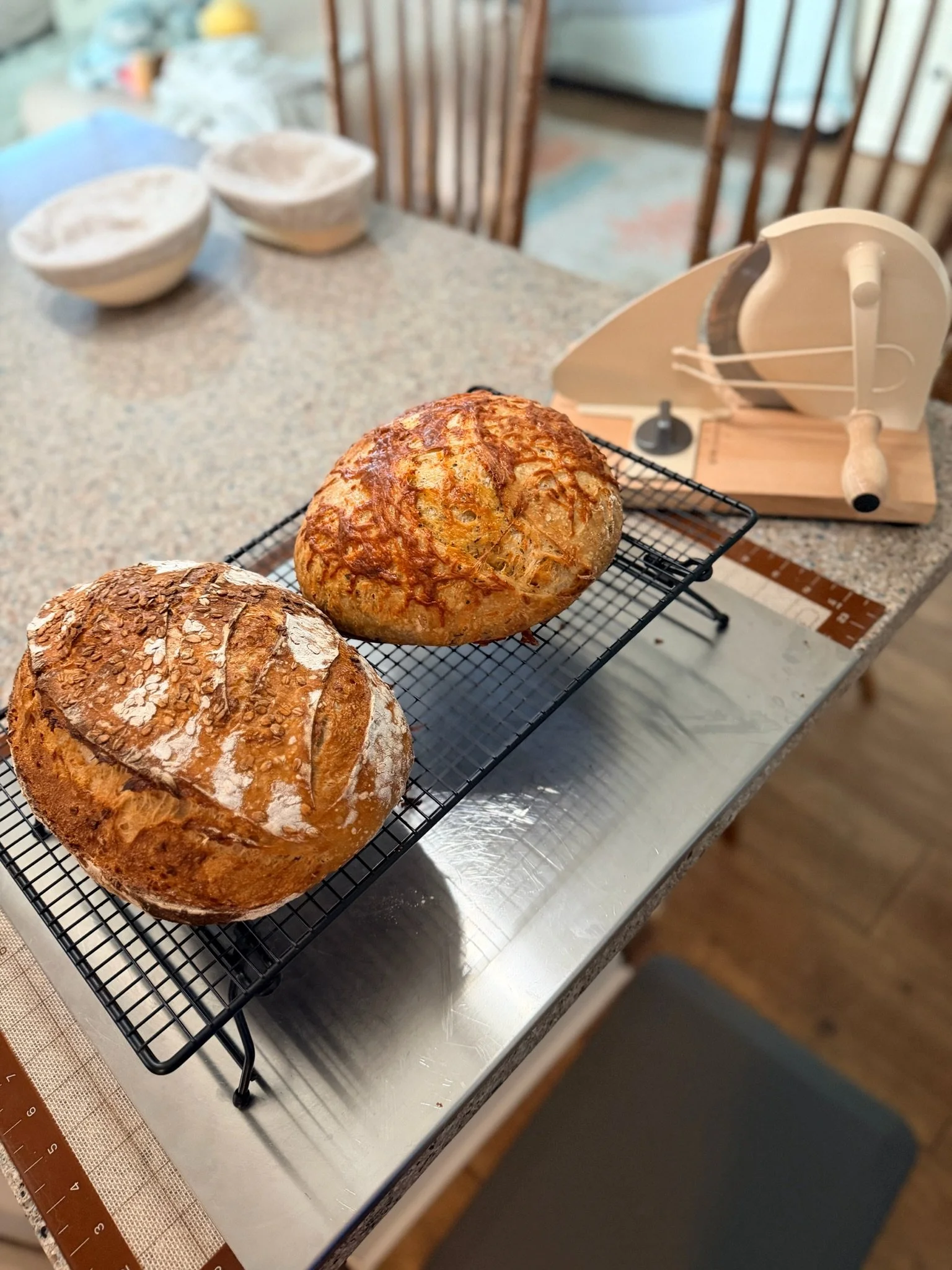 Three freshly baked bread loaves on a wire cooling rack on a kitchen counter with a bread slicing machine in the background.