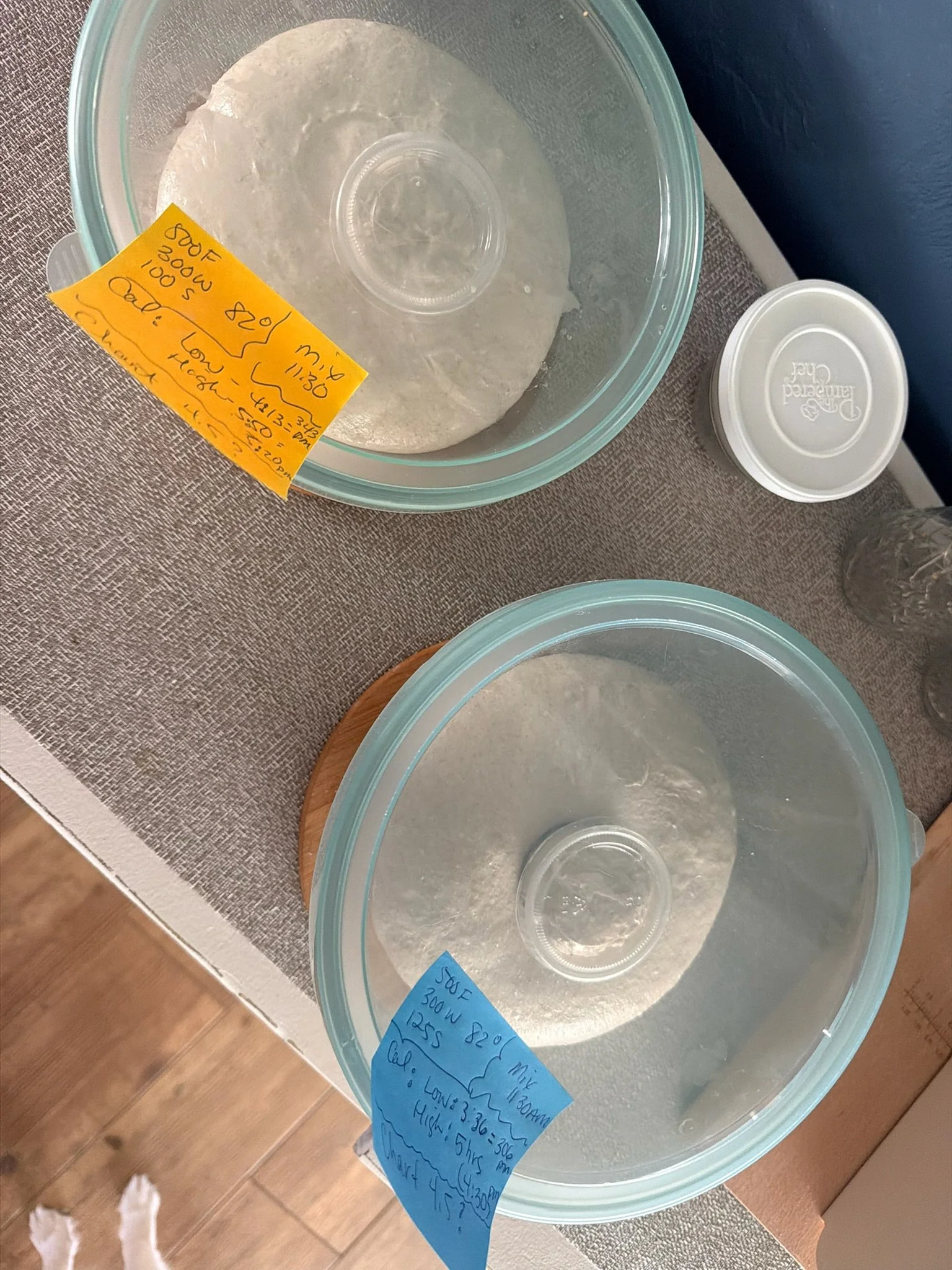 A clear plastic container with a lid, containing a piece of dough covered with plastic wrap, sitting on a table with a tan tablecloth. There are sticky notes with handwritten notes attached to the container.