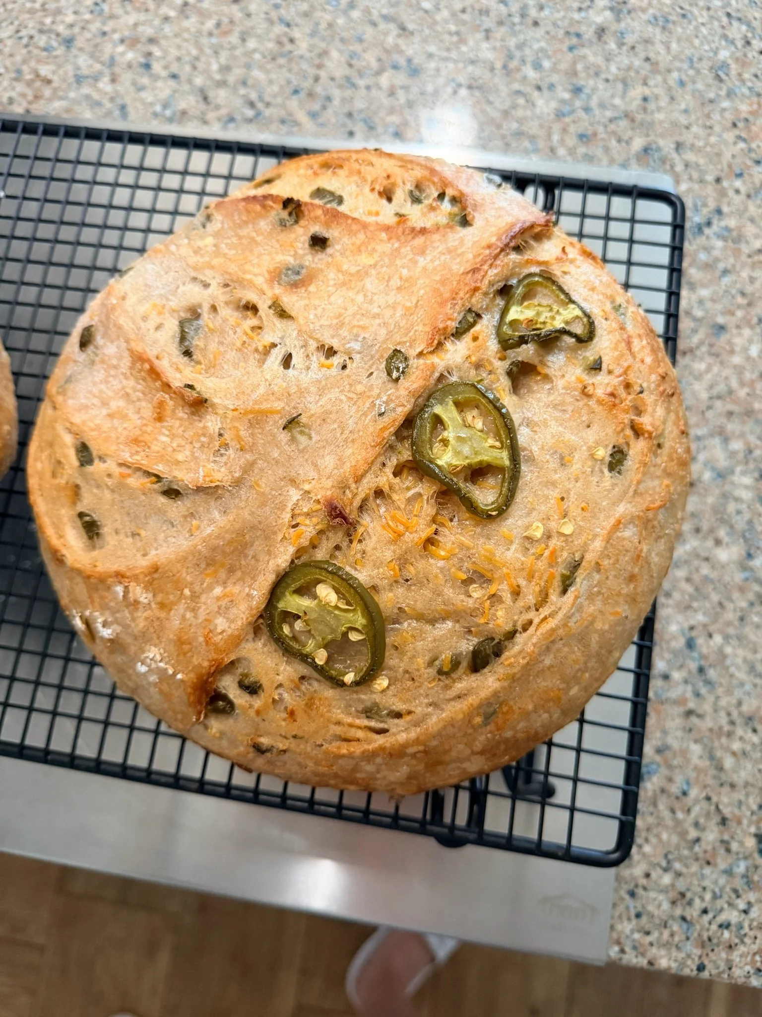 Close-up of a loaf of bread with jalapeno slices on top, resting on a wire cooling rack on a kitchen countertop.