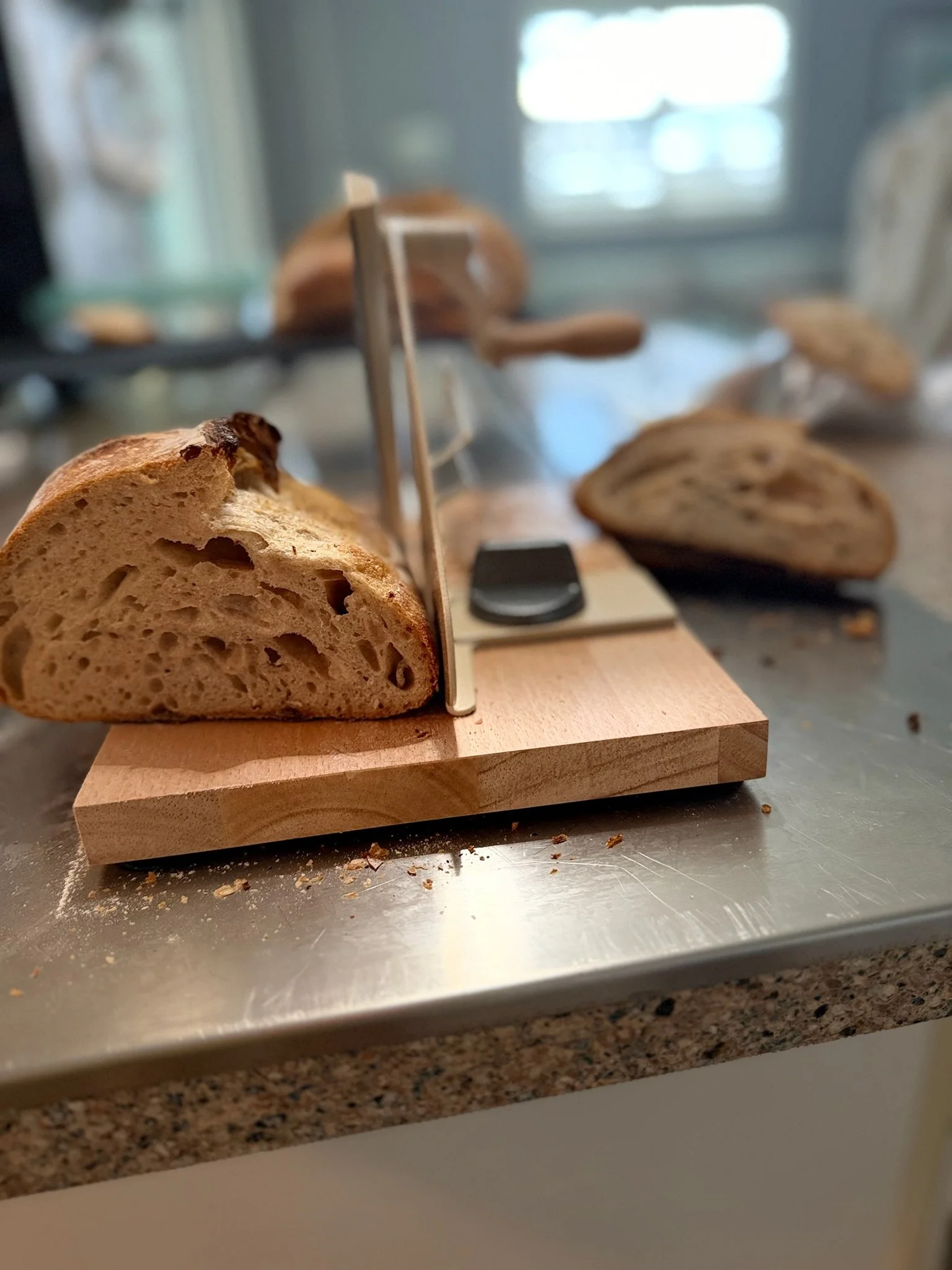Close-up of a slice of bread on a wooden cutting board with a bread knife, with other bread loaves in the background on a kitchen countertop.