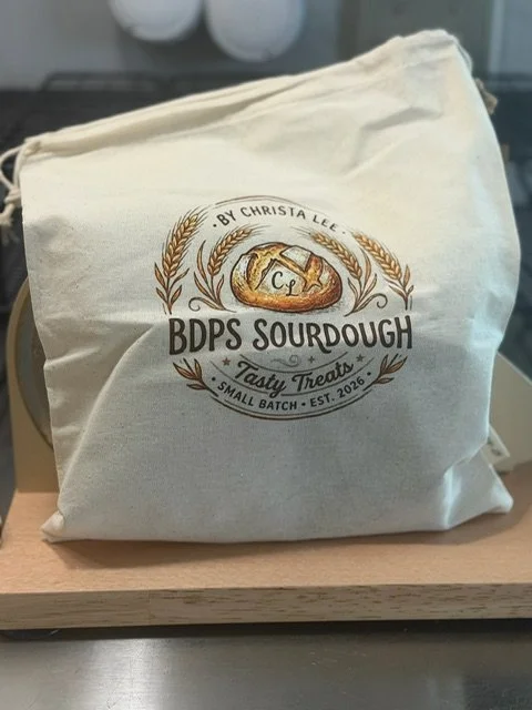 A white bag with a logo that says 'BDPS Sourdough, Tasty Treats, Small Batch, Est. 2026' and features an image of a sourdough bread with a stamped 'C+L' in the middle, surrounded by wheat images.