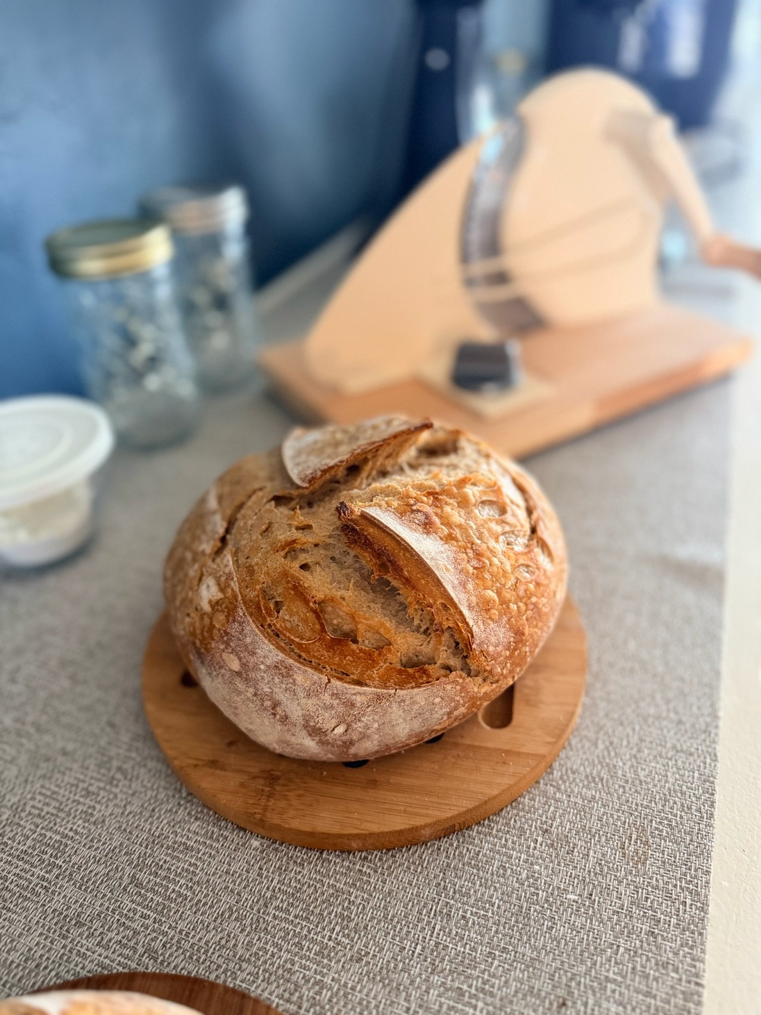A loaf of crusty bread resting on a round wooden cutting board on a textured tablecloth.