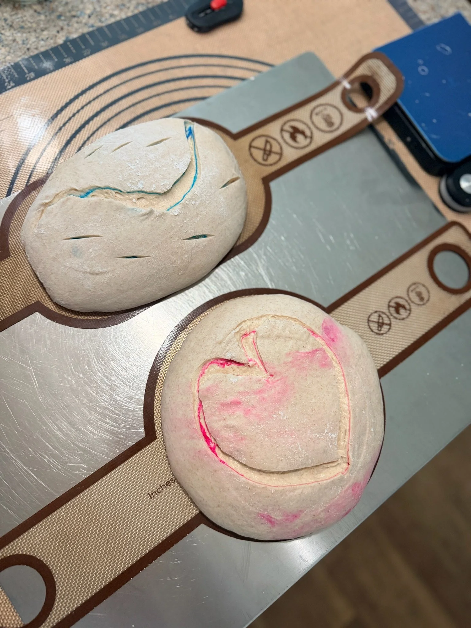 Photo of two bread dough balls on a silicone baking mat, with one ball scored with a blue marker and the other with pink. The mat has measurement markings and baking instructions.