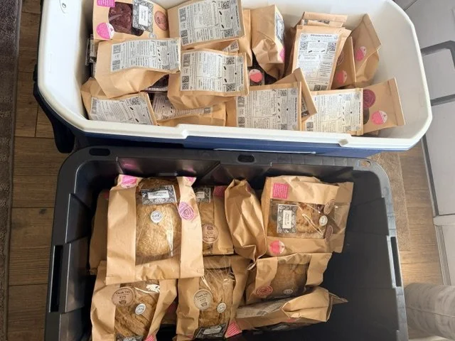 Two storage bins filled with several brown paper bags of products, some with clear windows showing contents, and labels or stickers. The bins are on a wooden floor.