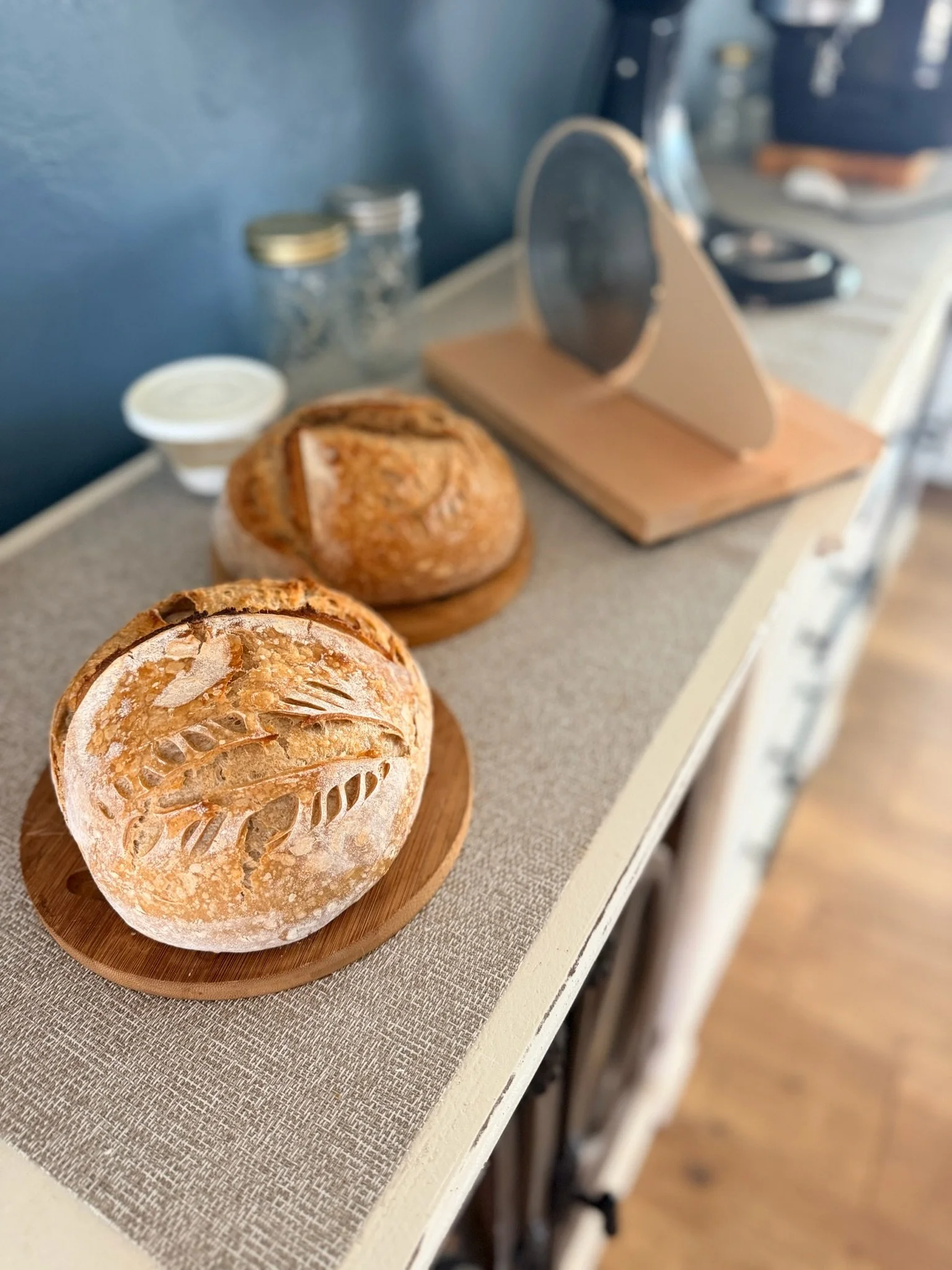 Close-up of artisan bread loaves on a wooden board, with jars of ingredients and kitchen utensils blurred in the background on a kitchen counter.