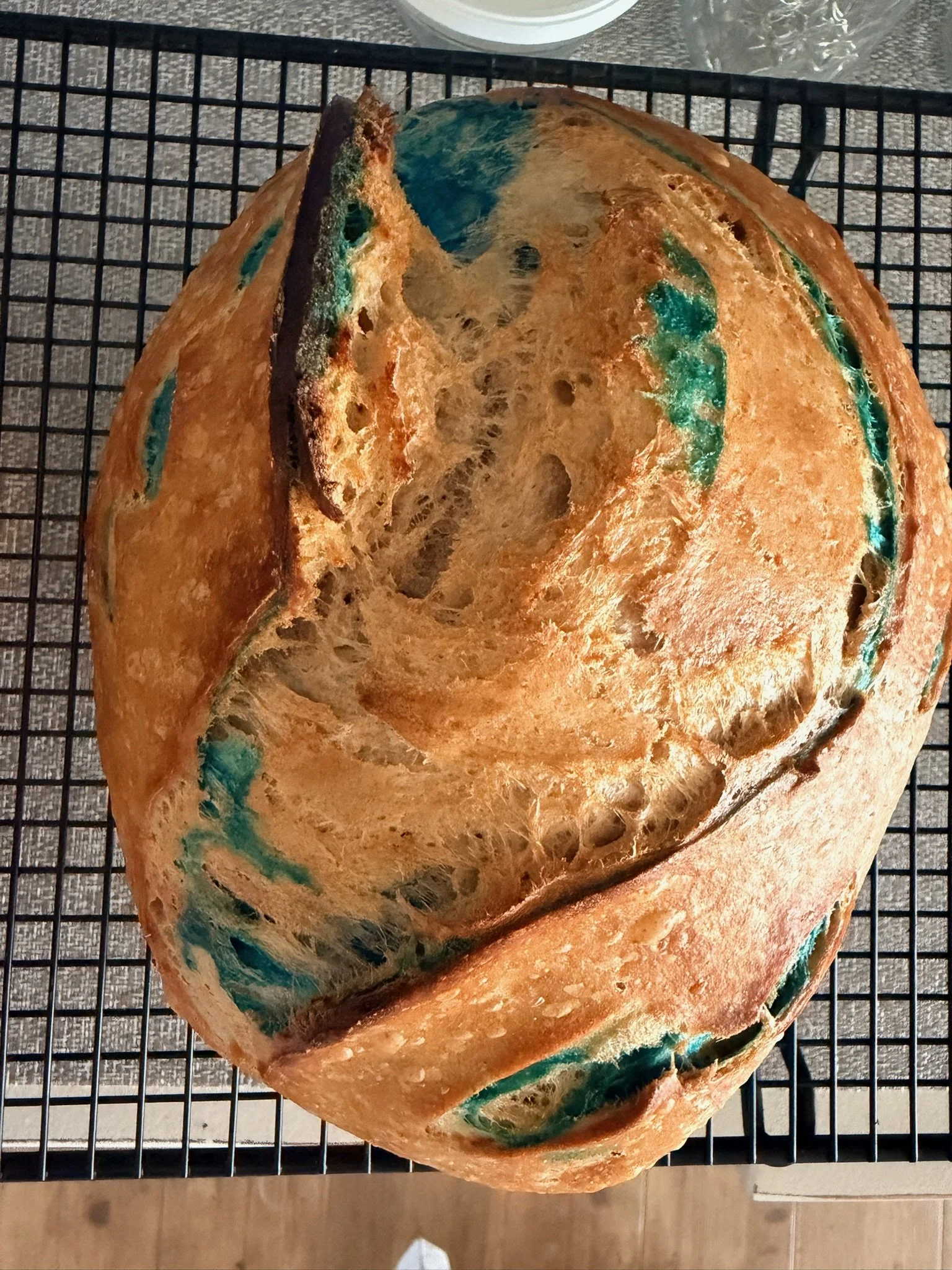 A loaf of bread with a crusty exterior, decorated with swirls of blue and green colored icing or glaze, cooling on a wire rack.