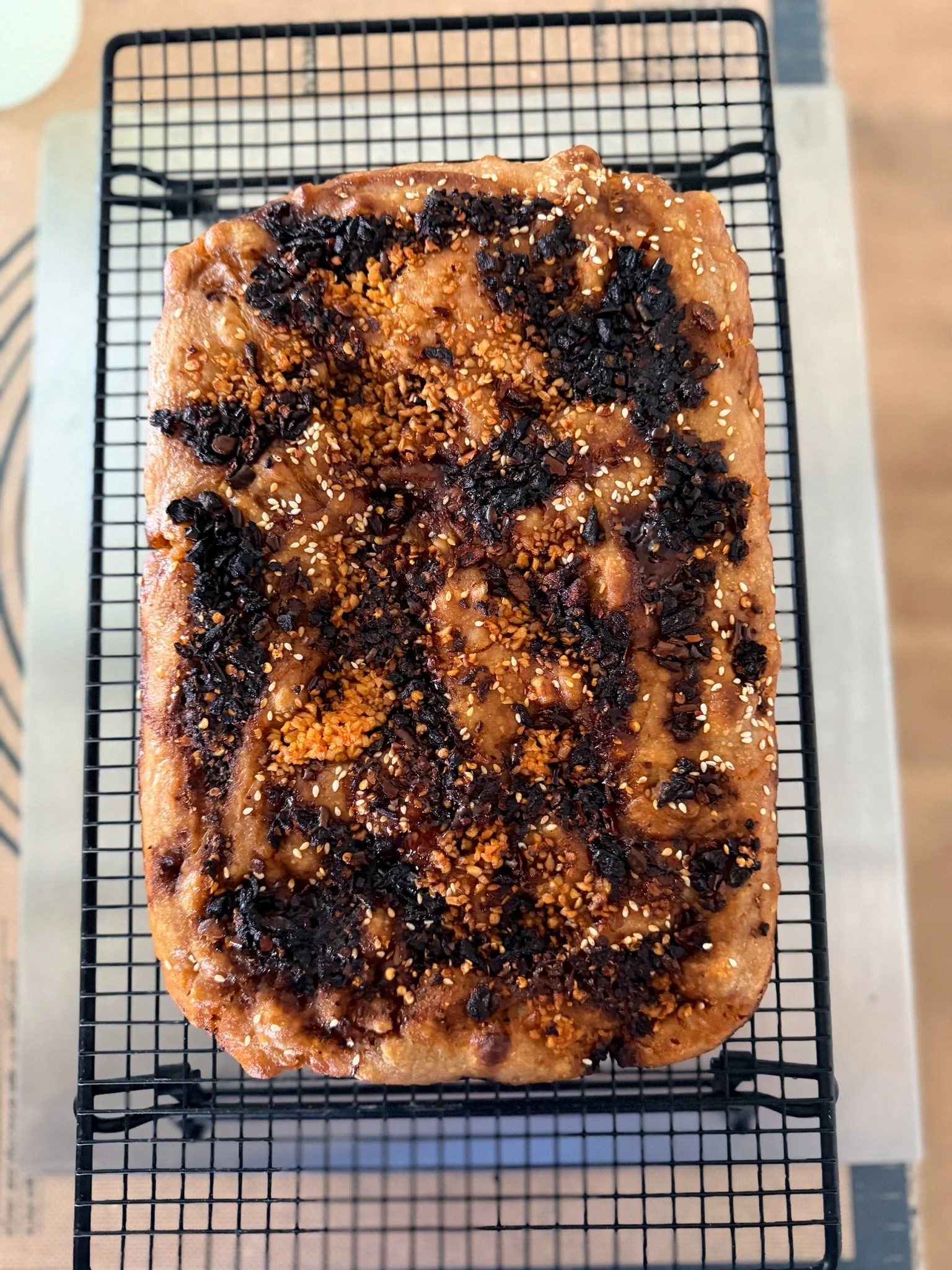 A rectangular cake with a golden-brown crust, topped with black and light brown crushed toppings and sprinkled with white sesame seeds, resting on a black wire cooling rack.