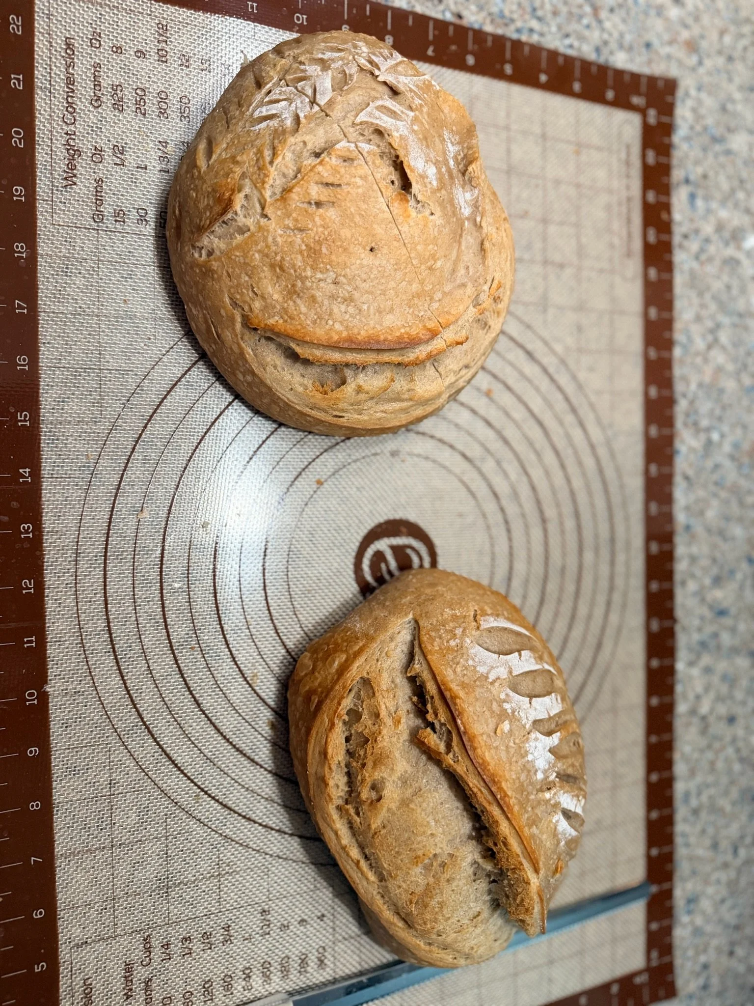 A round loaf of bread with a smiley face carved on top, placed on a silicone baking mat with measurement markings.