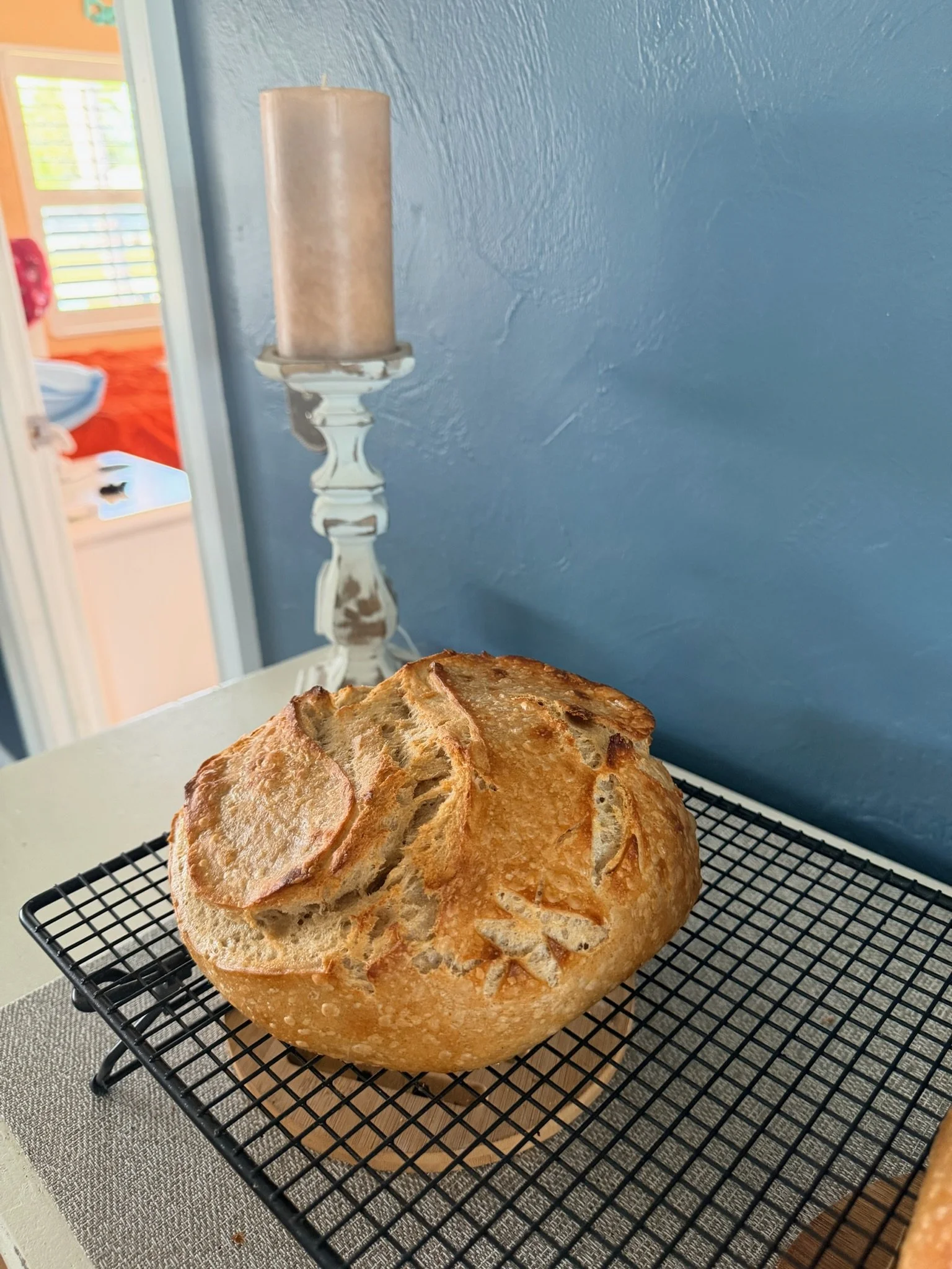 A freshly baked loaf of bread with a golden crust resting on a wire cooling rack in a kitchen. In the background, there is a candlestick with a large candle against a blue wall.