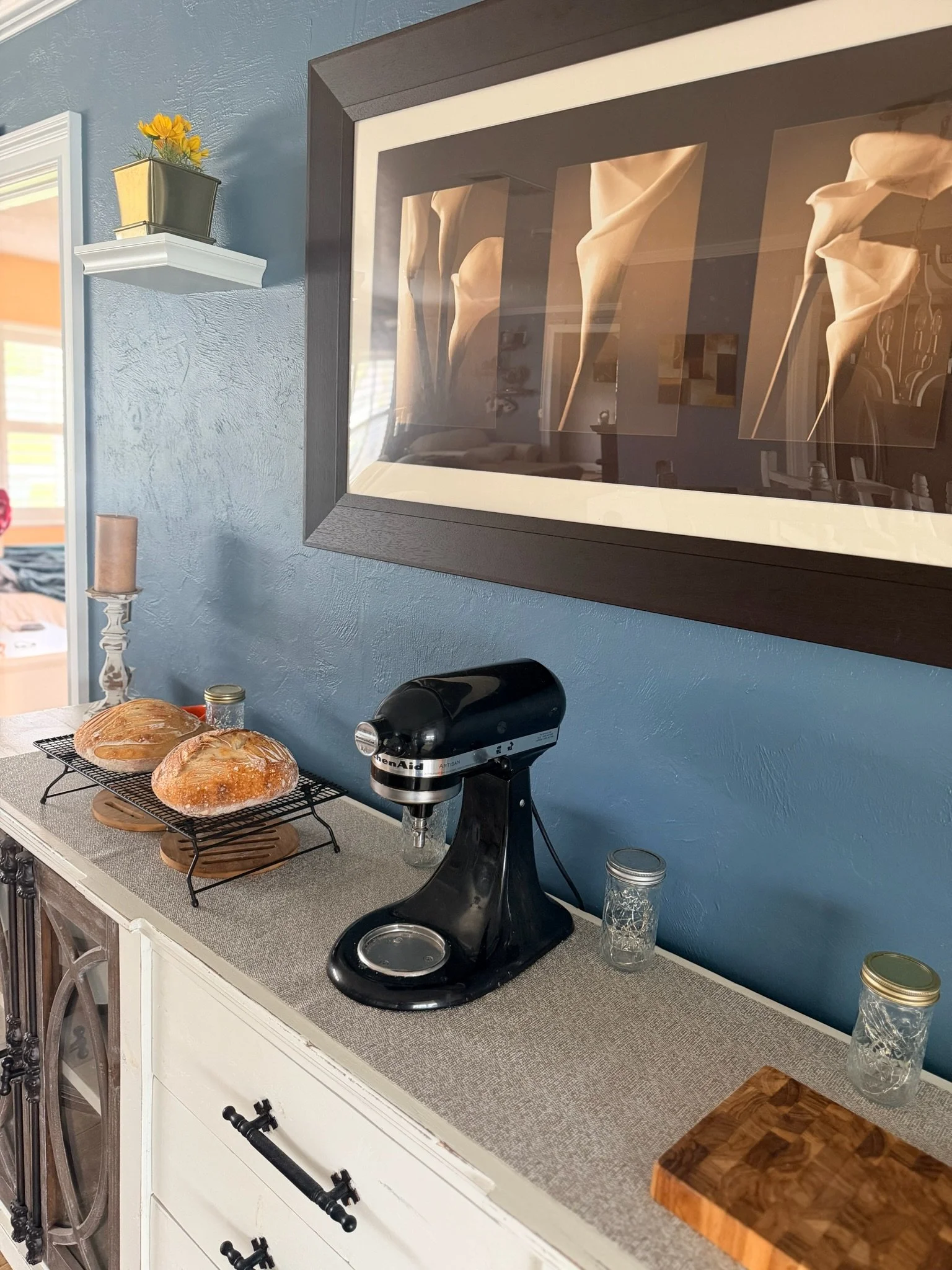 Kitchen countertop with two loaves of bread on a cooling rack, a stand mixer, and jars. A framed artwork of calla lilies hangs on the blue wall behind.