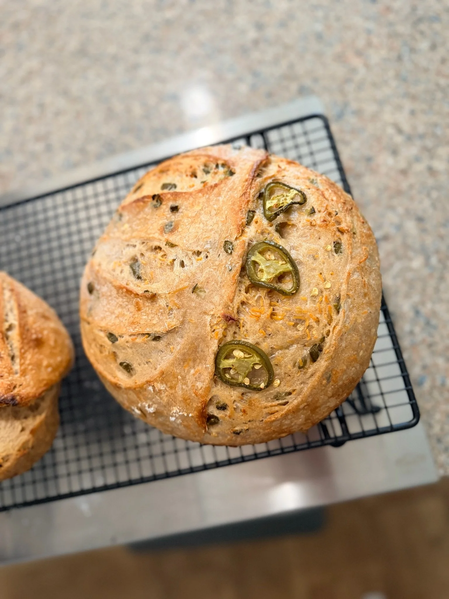 Round bread with jalapeño slices and herbs on top, resting on a cooling rack.