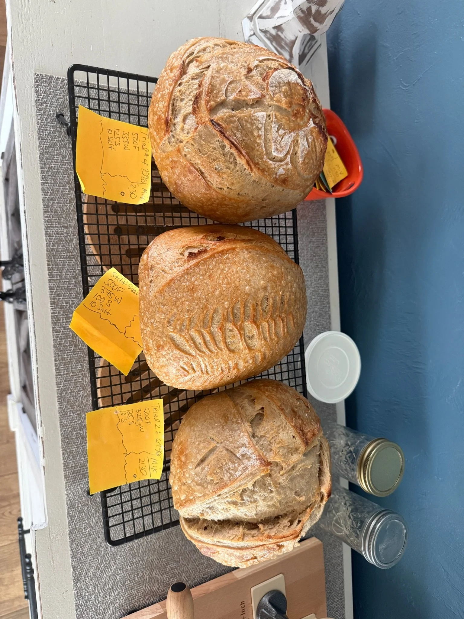Three loaves of bread on a cooling rack and countertop with jars and jars of bread mixes in the background.