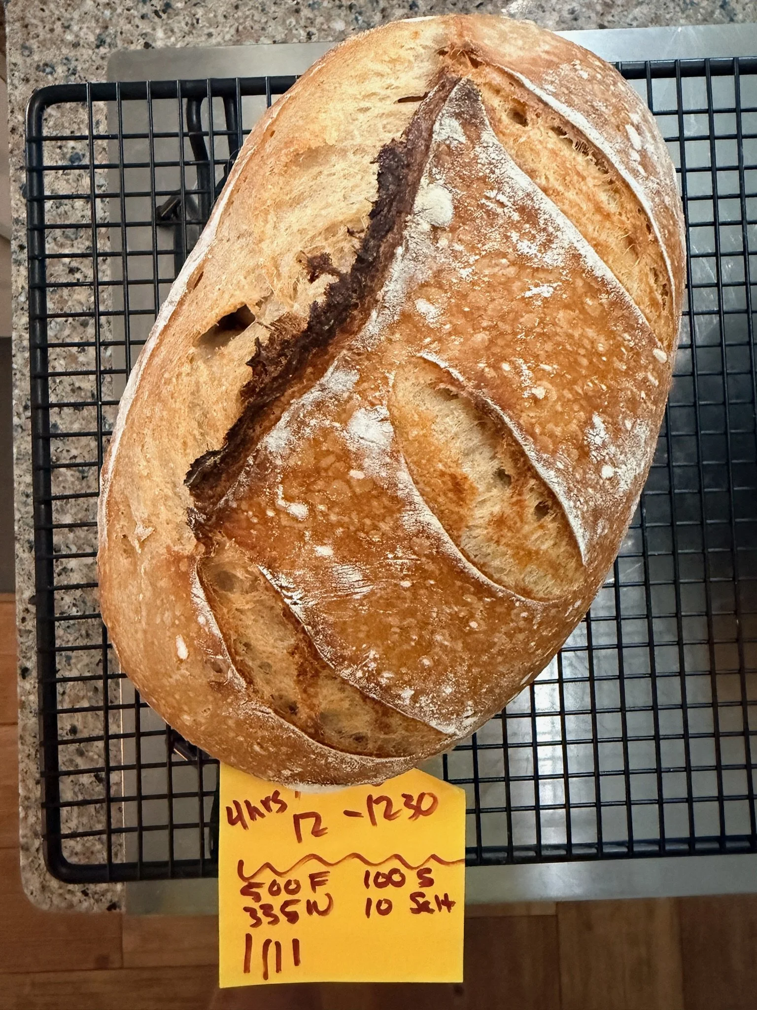 Large round loaf of bread on a cooling rack, with a dark crust and dusted with flour, next to a handwritten price sign.