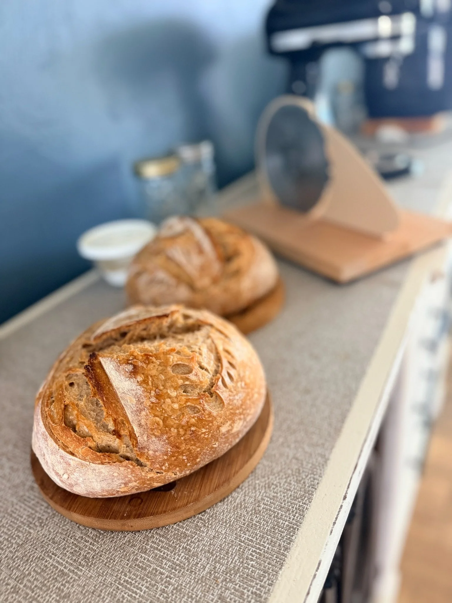 Fresh bread roll on a wooden board in the foreground, with a loaf and a jar of butter or cream behind it, on a table covered with a tablecloth.