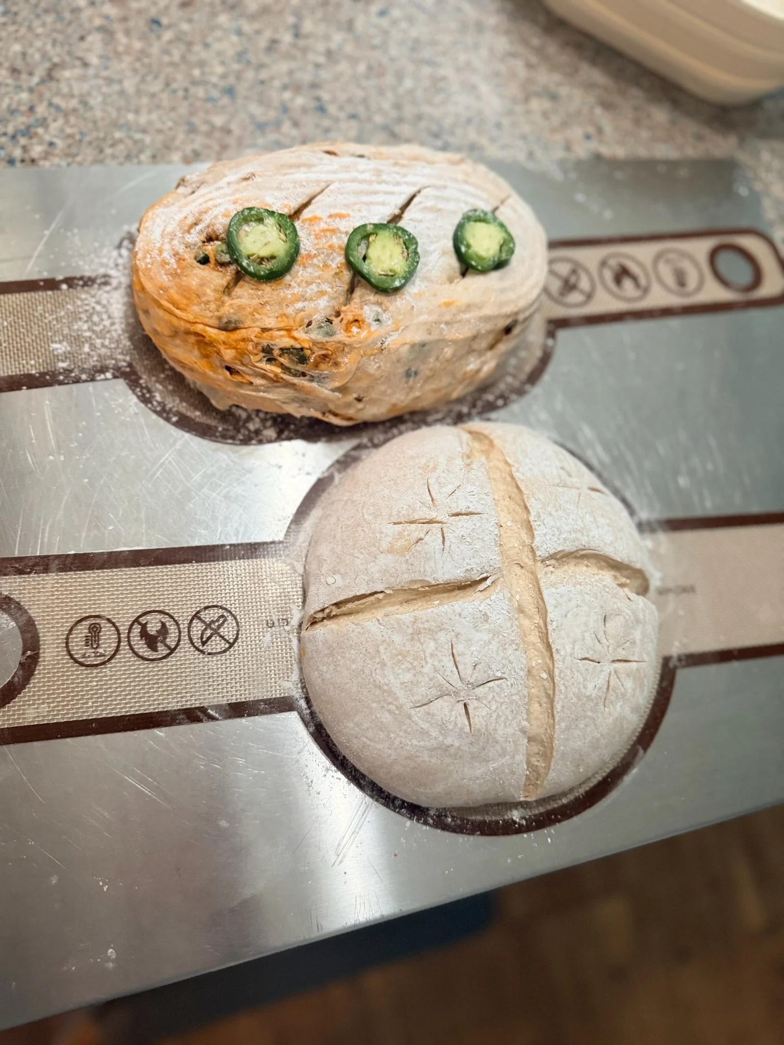 A loaf of bread with sliced jalapeño peppers on top, resting on a silpat baking mat with a second unbaked loaf nearby.