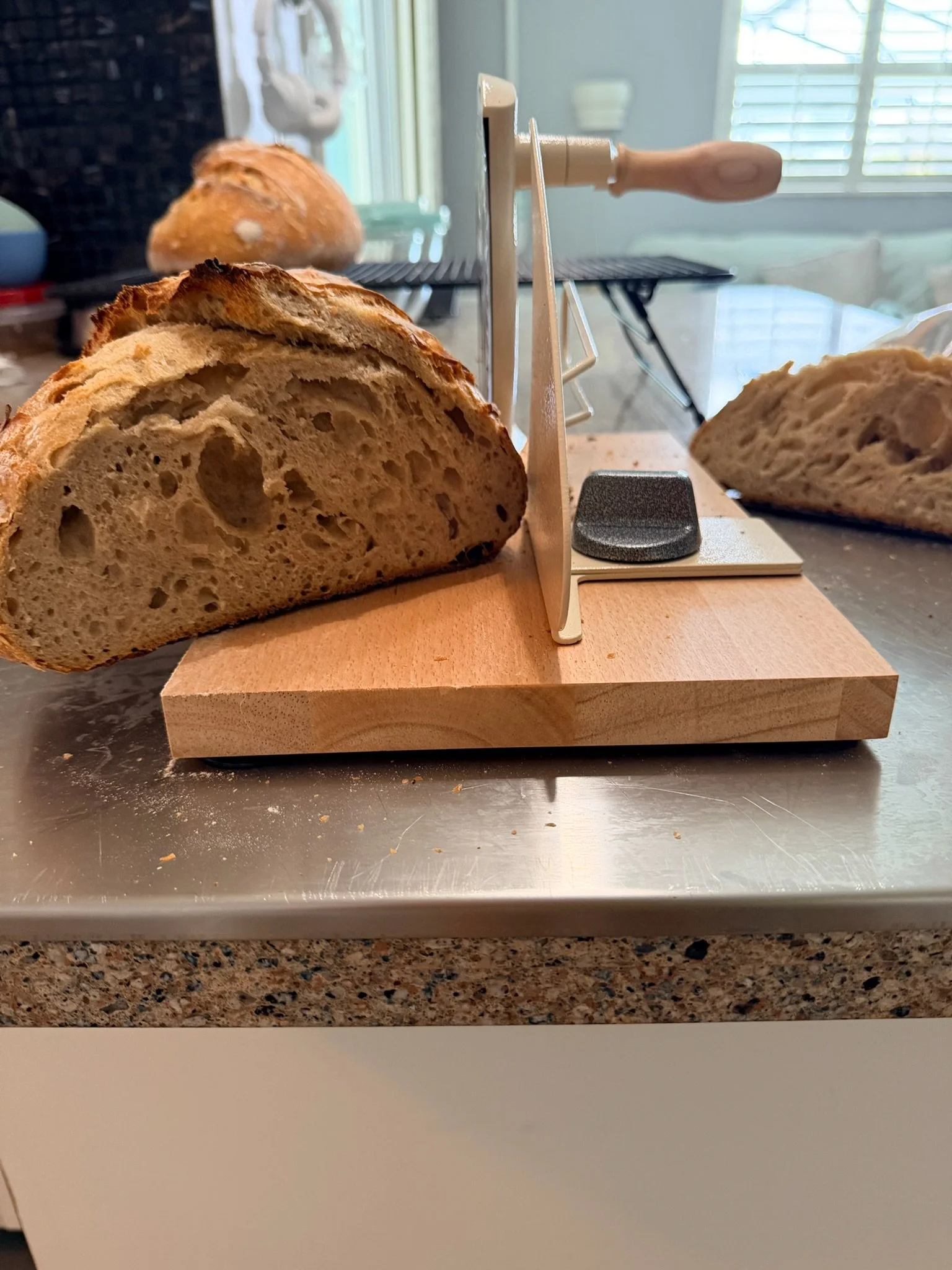 Sliced loaf of bread on a wooden cutting board with a bread slicing guide, with more bread in the background near a wire cooling rack, in a kitchen with natural light coming through the window.