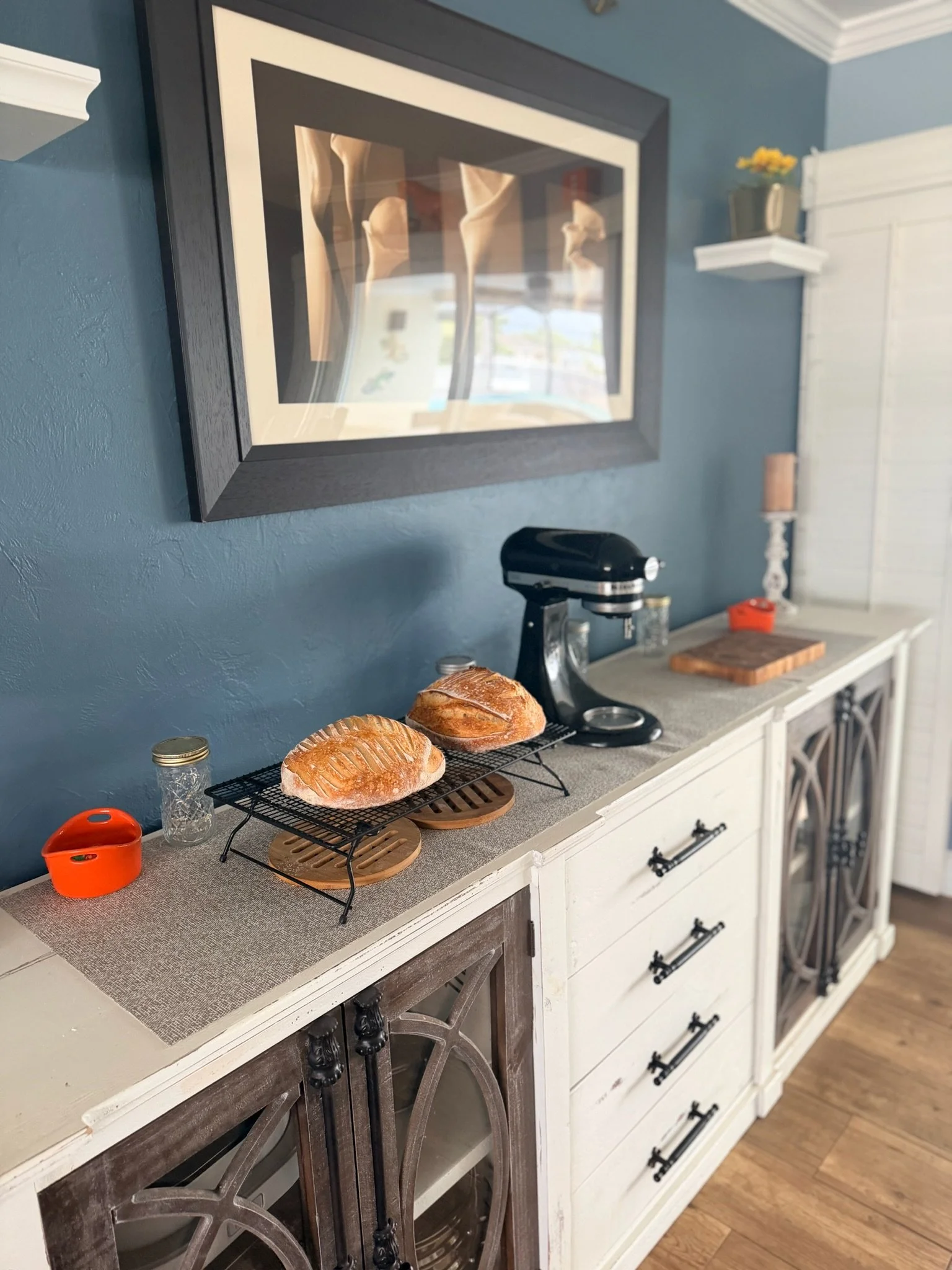 A white kitchen sideboard with black handles, two sections with glass doors, and a butcher block on top. Next to the sideboard is a white cabinet with a shelf holding a potted plant with yellow flowers. On the countertop are a black mixer, a glass ja