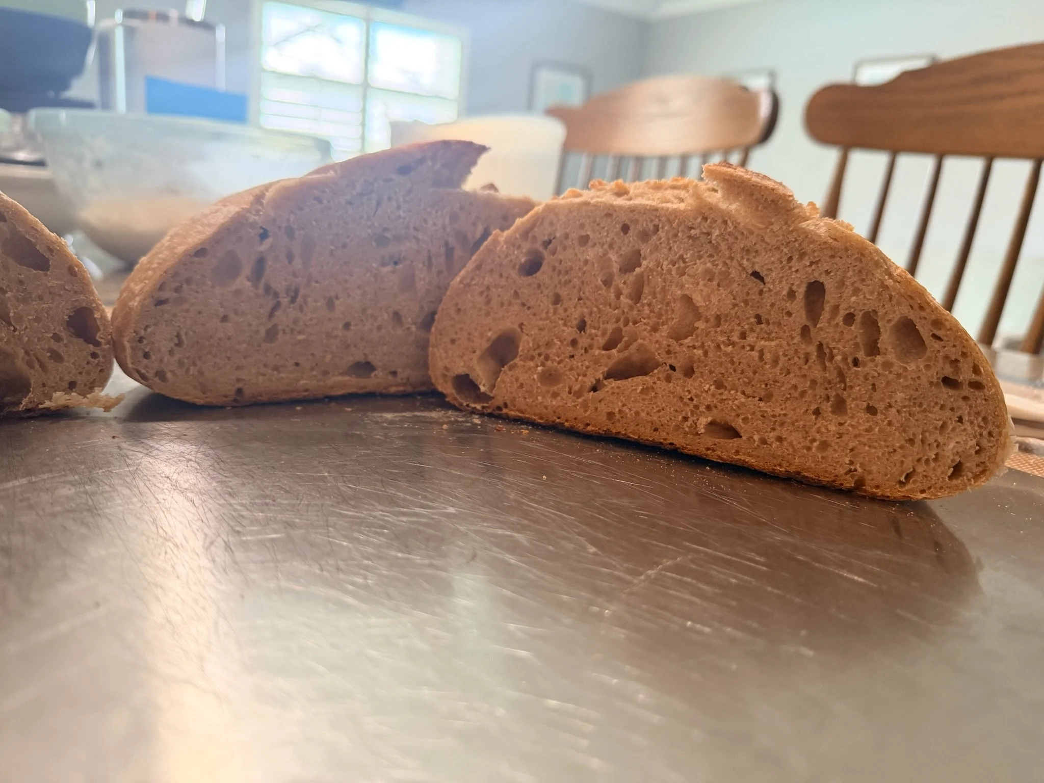 Close-up of sliced bread on a metallic surface in a kitchen setting with chairs and a window in the background.