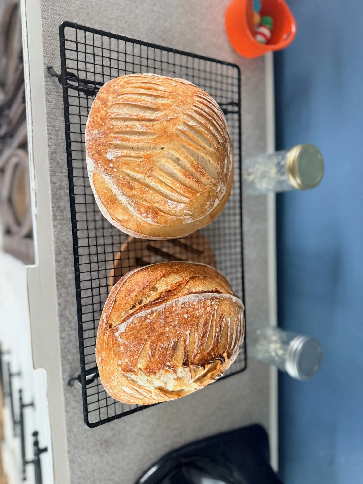 Two freshly baked bread rolls on a black wire rack on a gray countertop, with a red container holding colorful candies and a glass bottle with a metal cap nearby.