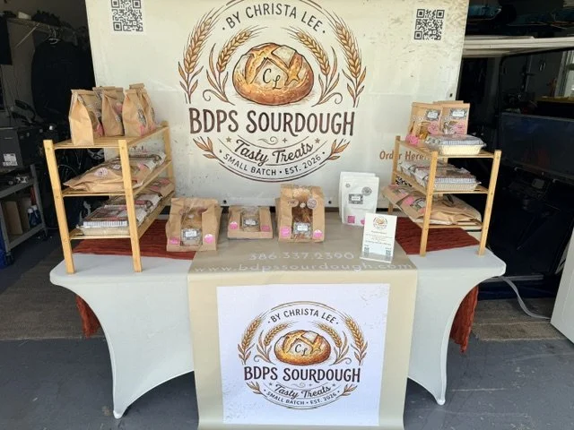 Display table for BDPS Sourdough by Christa Lee with baked bread products on wooden shelves and a banner showing the brand logo, products, and contact information.