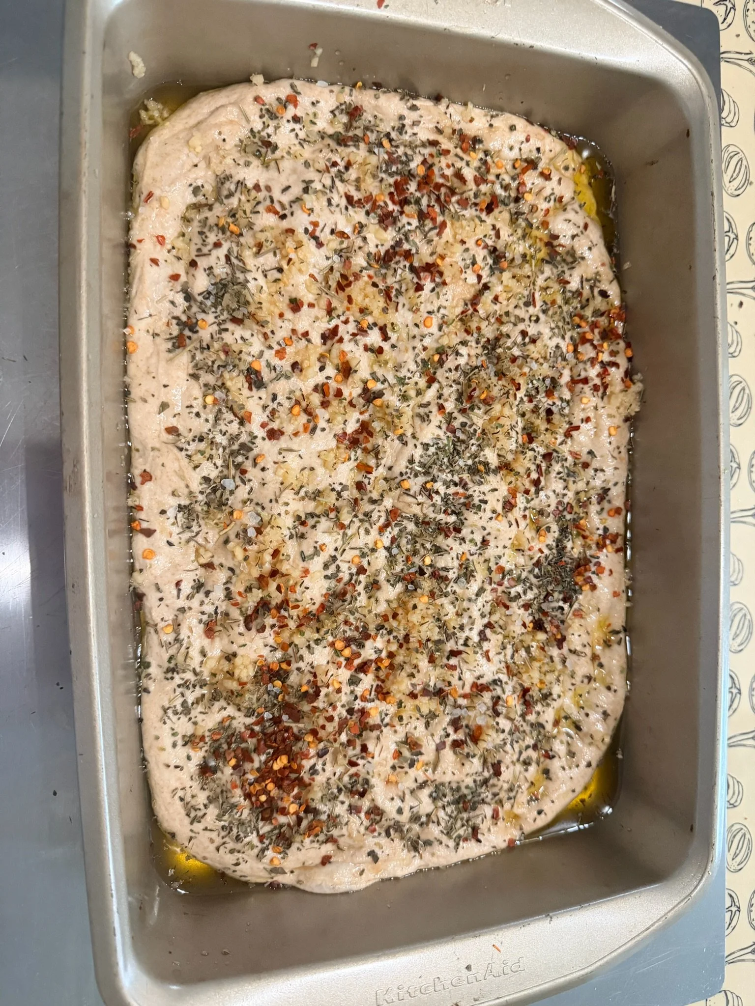 Raw chicken breast sprinkled with chopped herbs and red pepper flakes in a roasting pan with olive oil.
