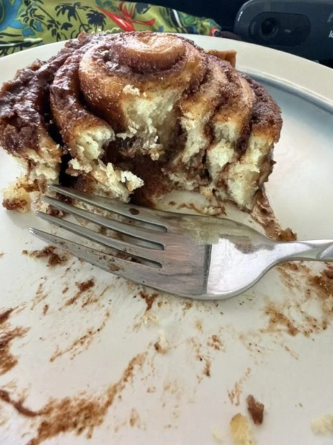 A partially eaten cinnamon roll with icing on a white plate, a fork nearby with some smudges, and a colorful cloth in the background.