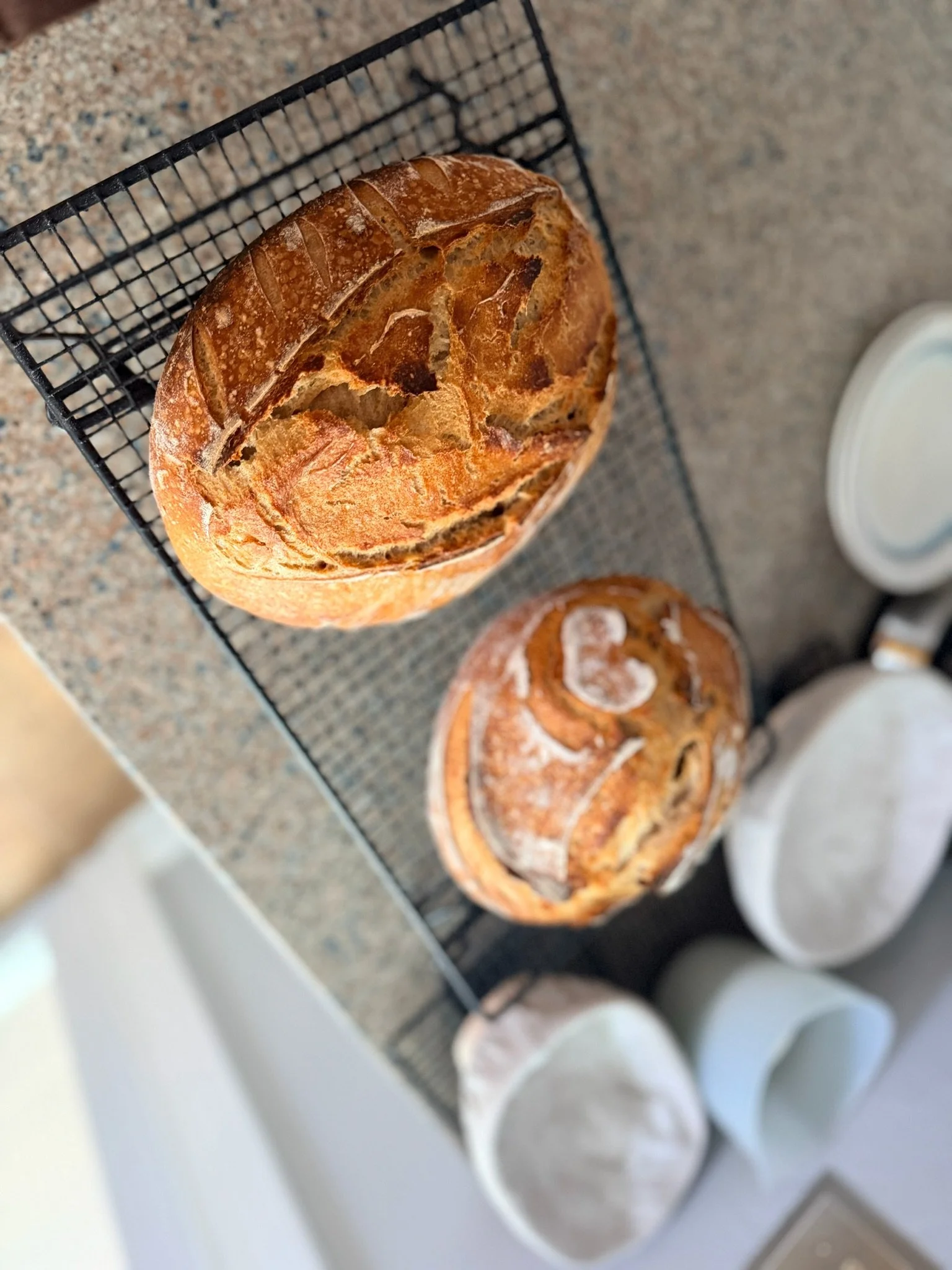 A loaf of bread resting on a wire cooling rack on a speckled countertop, with additional baked goods and white cups visible in the background.