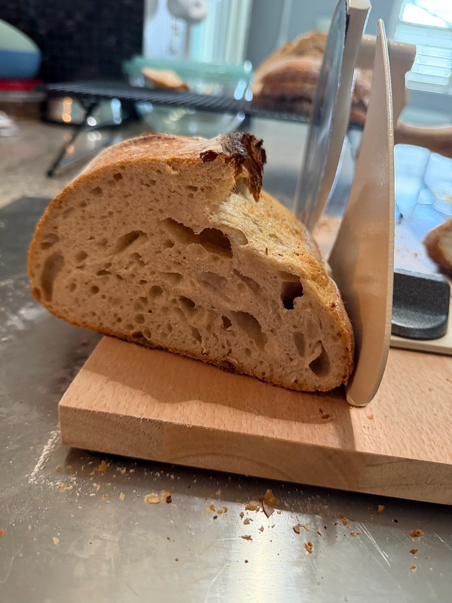 A slice of toasted bread with a crusty edge and a hole in the middle, resting on a wooden cutting board, with crumbs scattered on a metal surface.