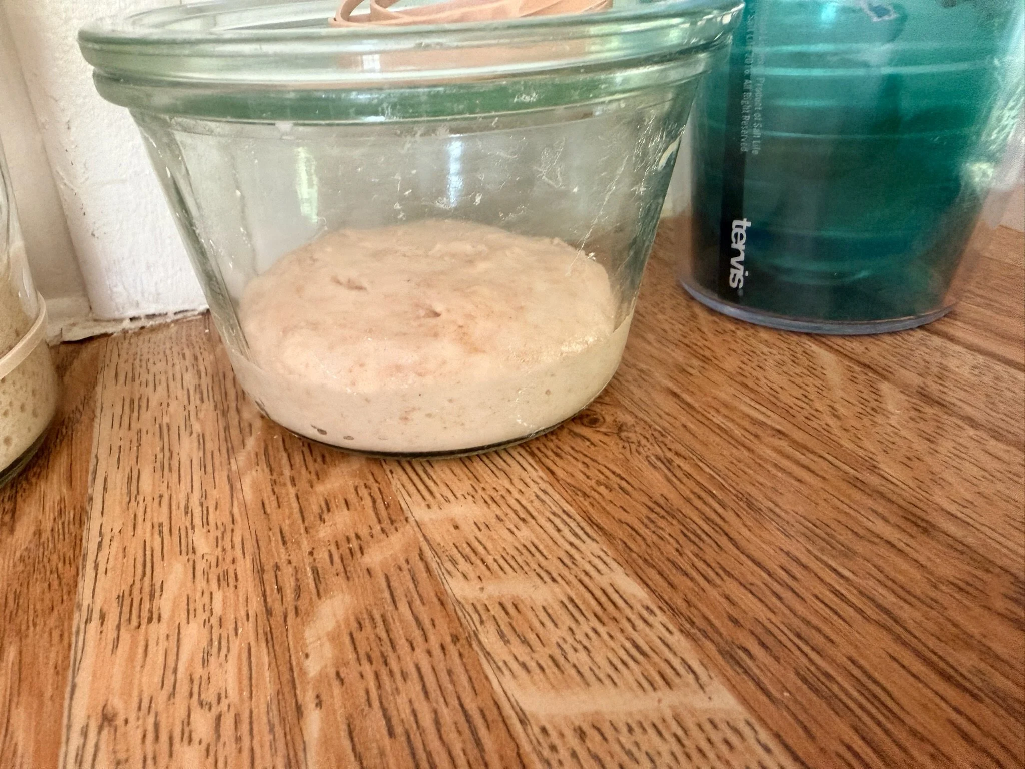 Close-up of a small glass bowl containing a beige, powdery substance, placed on a wooden surface, with a larger green container partially visible beside it.