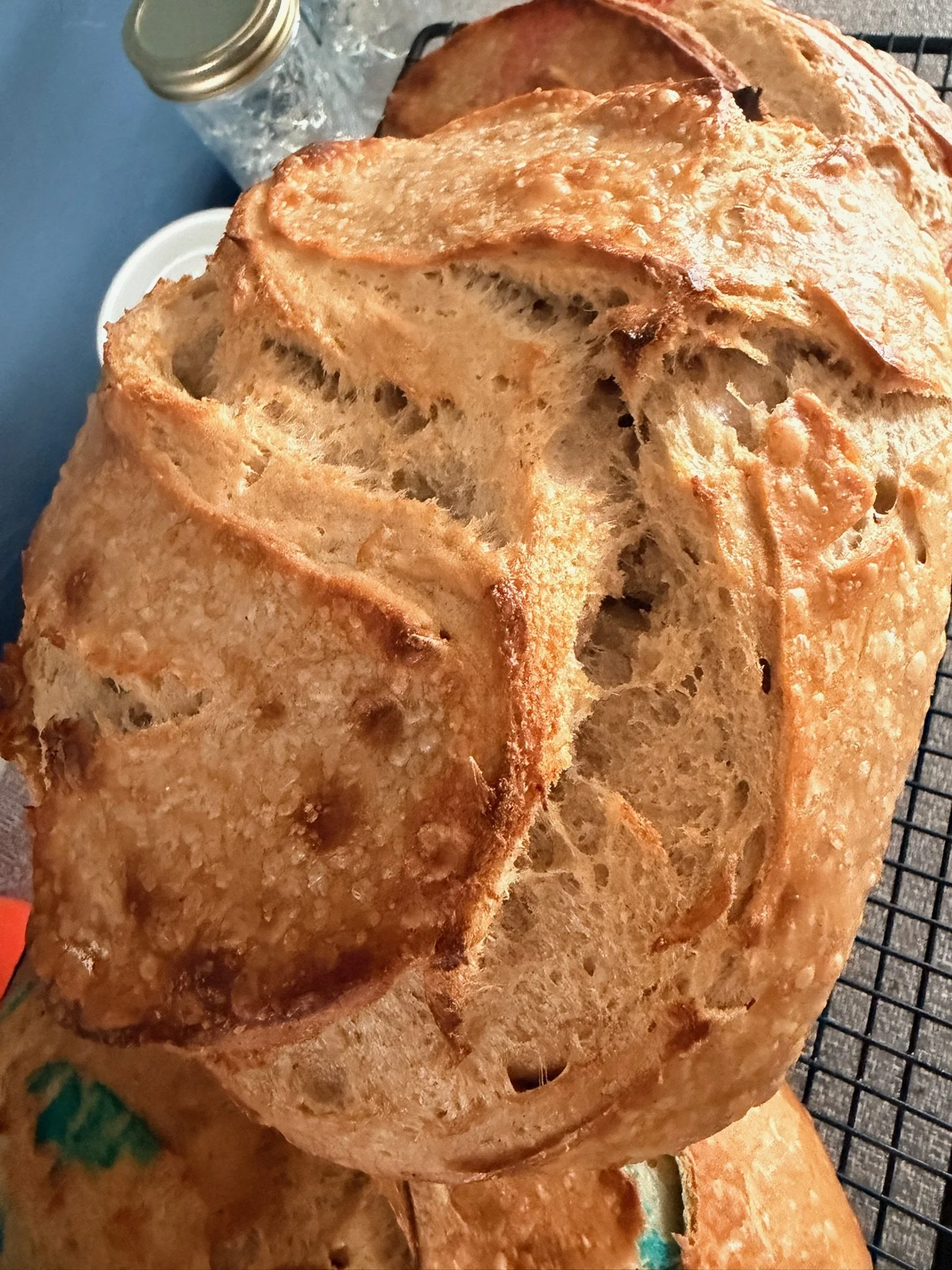 Close-up of a freshly baked loaf of bread with a golden-brown crust resting on a cooling rack. The bread appears to have a rustic, artisanal style with a textured surface.