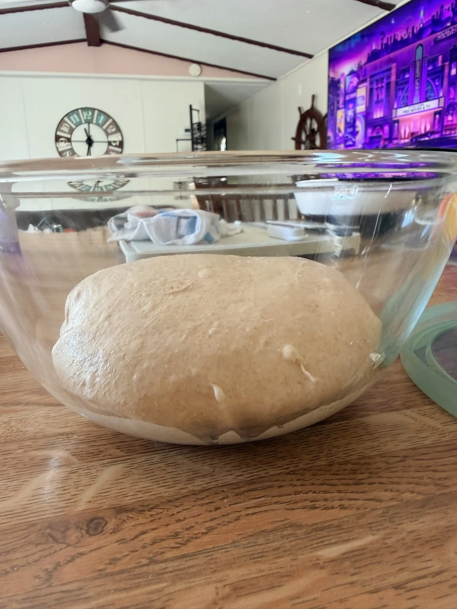 A glass bowl containing dough on a wooden surface, with a kitchen and a large TV in the background.
