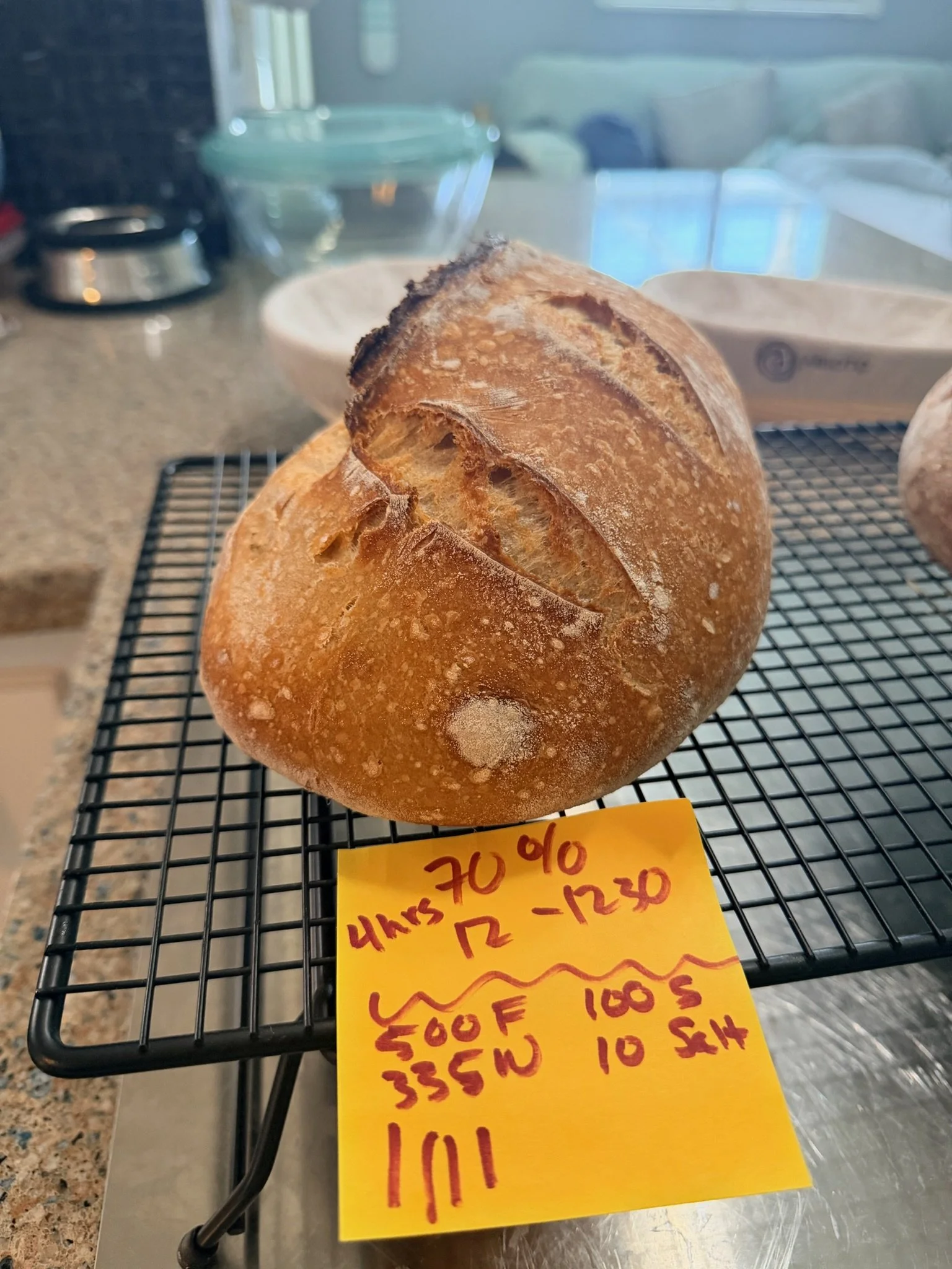 A loaf of bread on a cooling rack, with a yellow price tag and handwritten notes attached, in a kitchen setting.