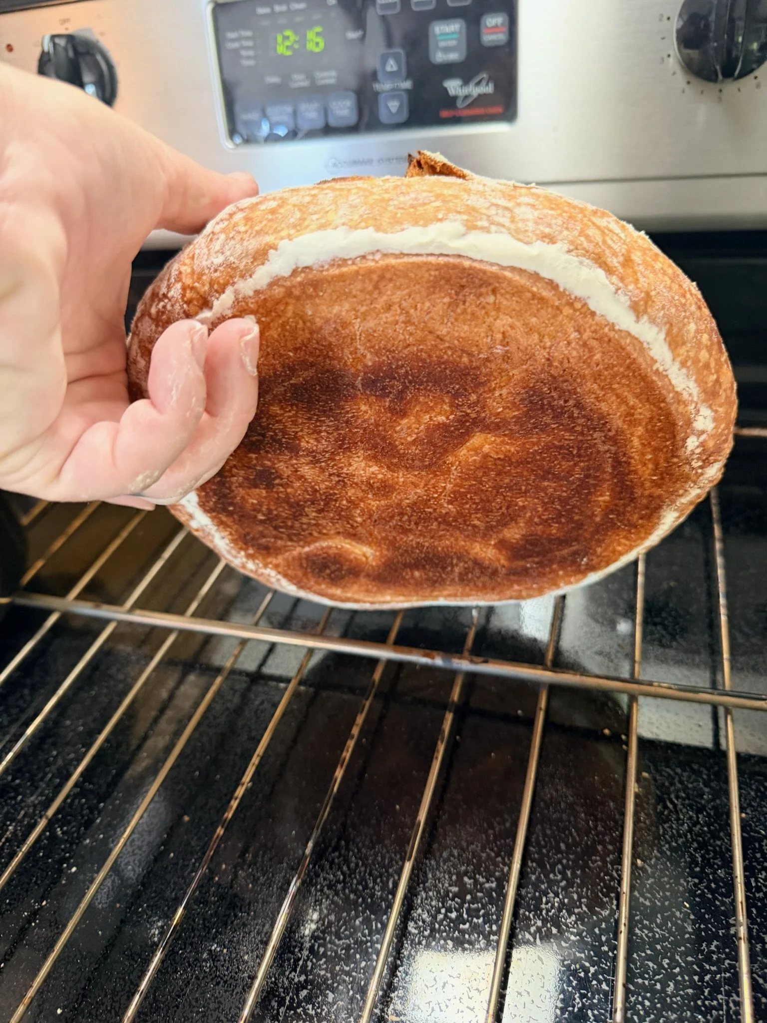A person is holding a toasted hamburger bun with a slice of cheese inside. The bun has a golden-brown toasted surface, and the person’s fingers are slightly dusted with flour. The oven temperature display shows 12:6.