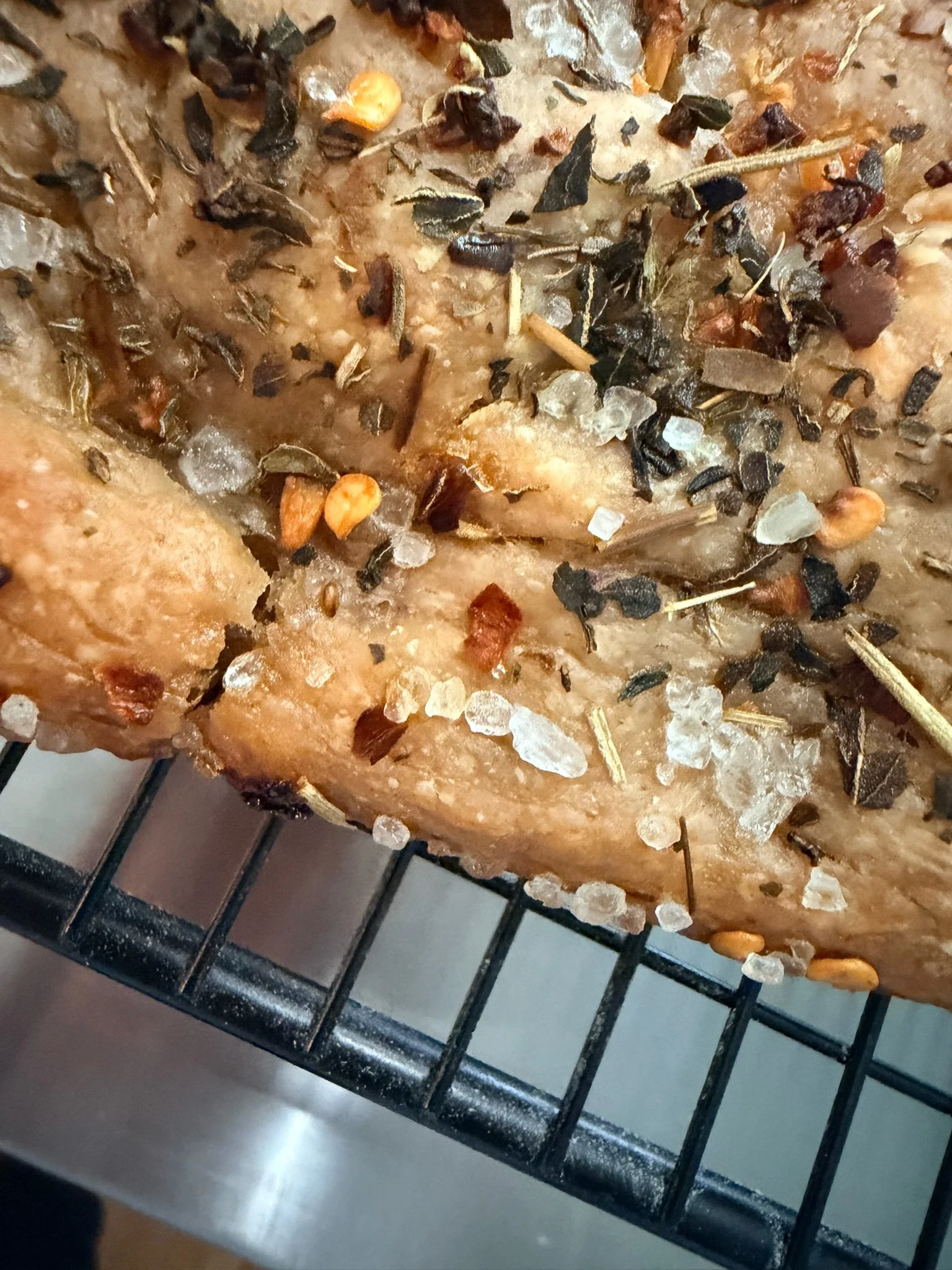 Close-up of a baked dish topped with various seasonings such as salt, pepper, and dried herbs, on a metal oven rack.