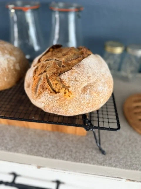 Close-up of a rustic round bread loaf with a crusty top, resting on a small black wire cooling rack in a kitchen.