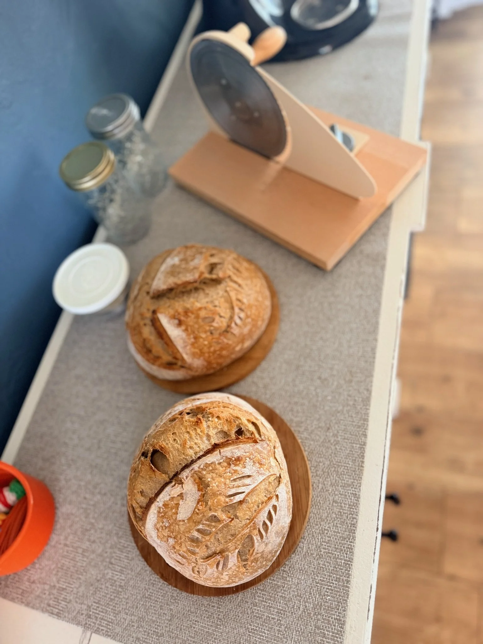 Three loaves of rustic bread on a wooden platter, with two glass jars and a napkin on a gray countertop.